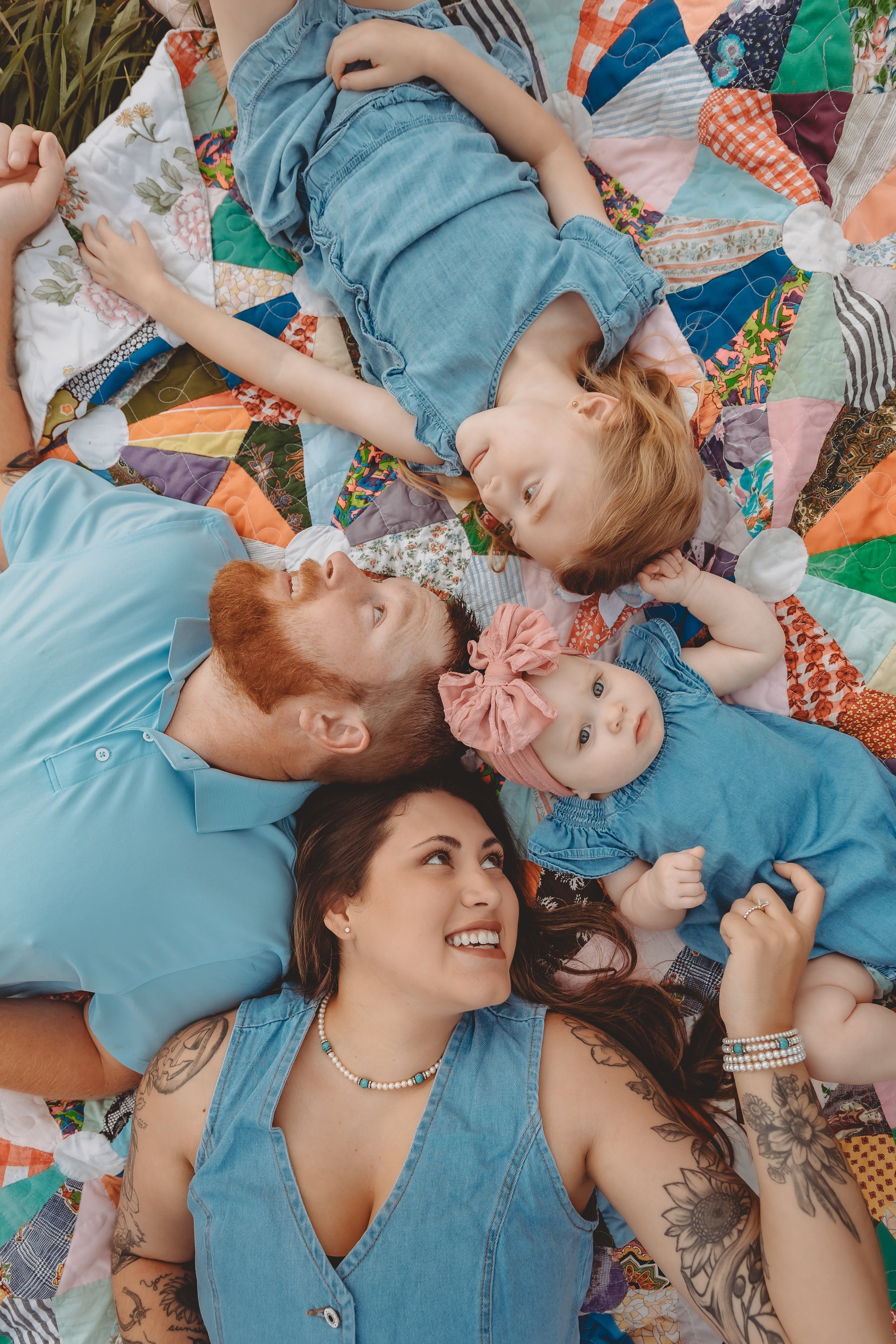 A woman and a man lying on a colorful patchwork quilt with two children, one girl and one baby, all relaxing and looking around.