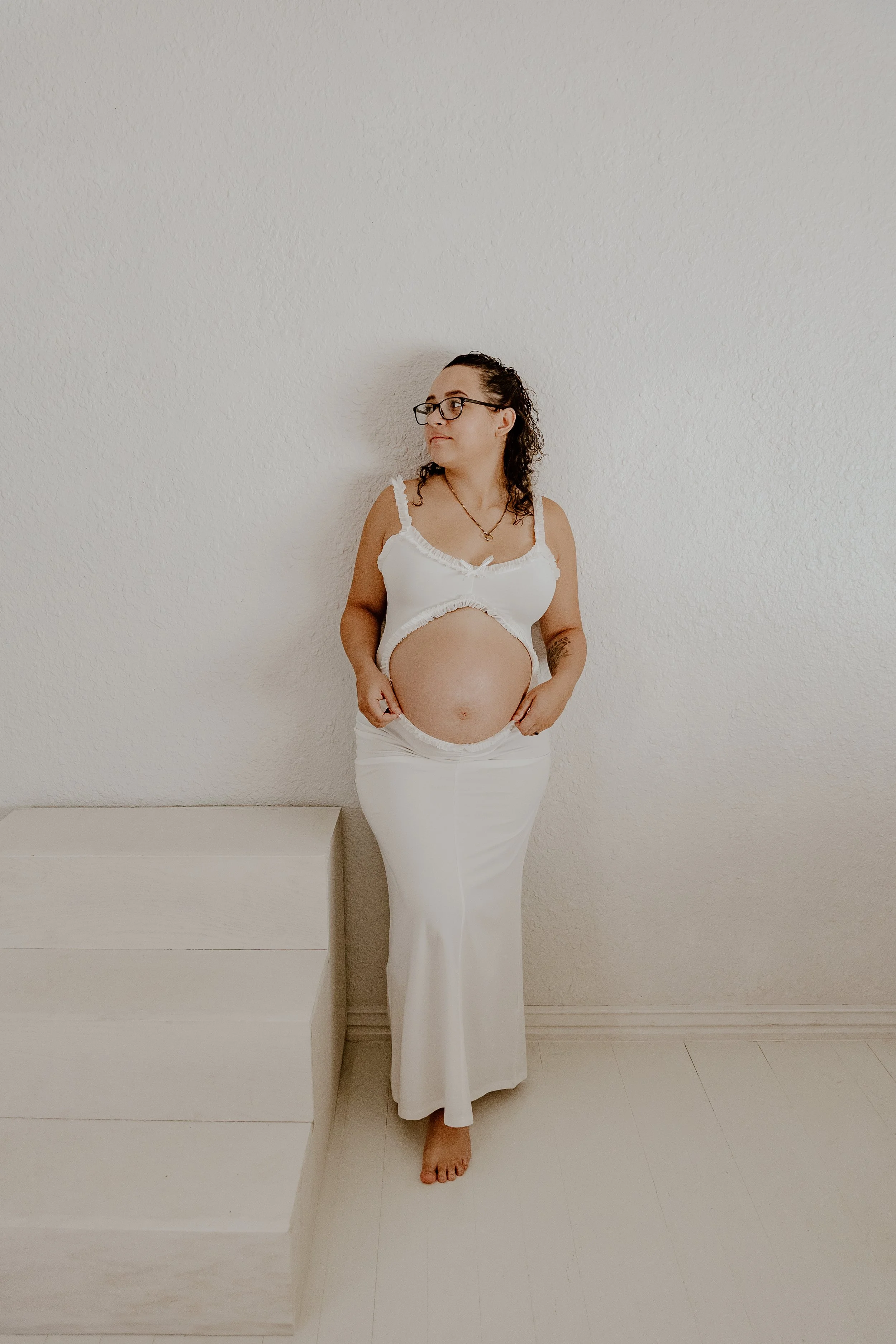 Pregnant woman with glasses in a white outfit standing by a staircase against a plain white wall. Studio 512 Fort Smith, AR Maternity photographer does a studio session.