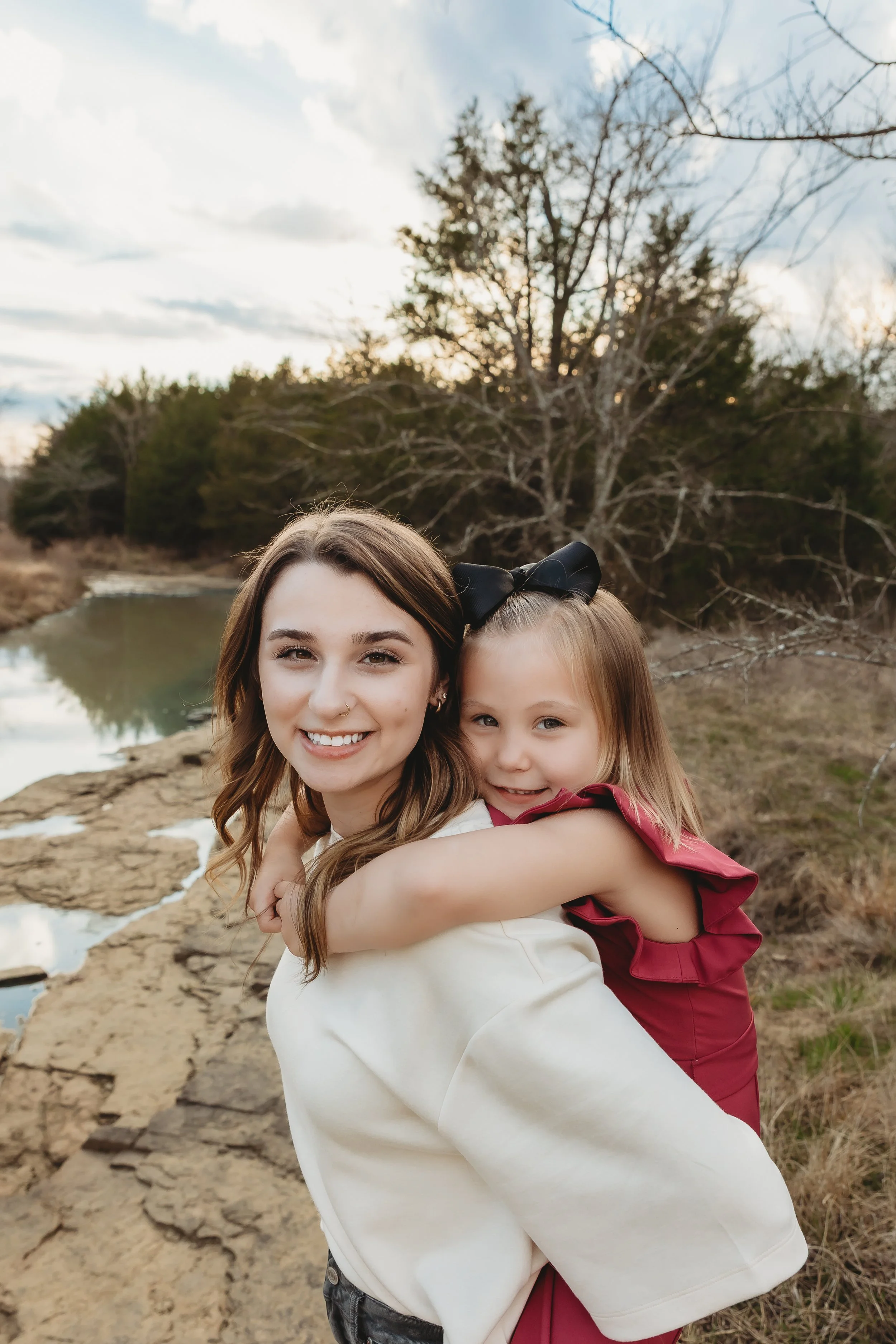 Two young women, one with long brown hair and a black bow, and the other with blonde hair, smiling and hugging outdoors near a creek with trees in the background.