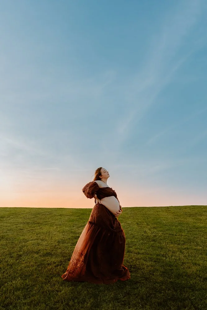 Pregnant woman in a brown dress standing on grass during sunset, holding her belly and looking up. Fort Smith, AR maternity photographer.