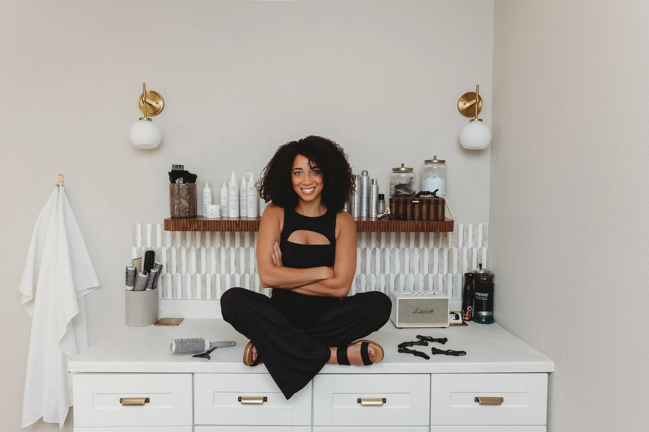 Woman sitting cross-legged on a white vanity table, surrounded by hair styling tools and products, with a neutral-colored wall and wall-mounted lights in the background.