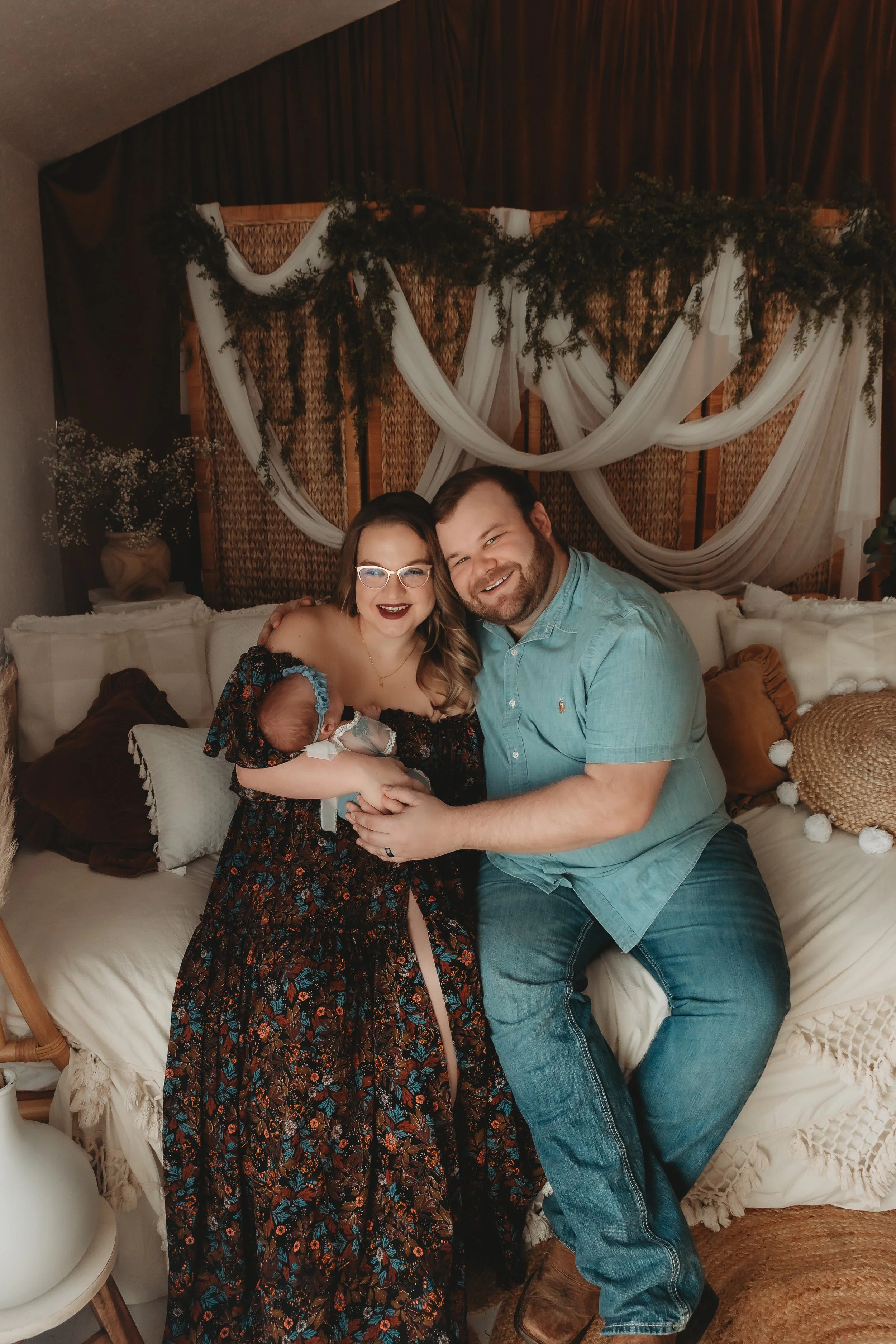 A family of three, including a woman, a man, and a baby, posing together on a bed in a warmly decorated room with draped fabric and greenery in the background. Newborn Photographer in Fort Smith, AR at studio location.
