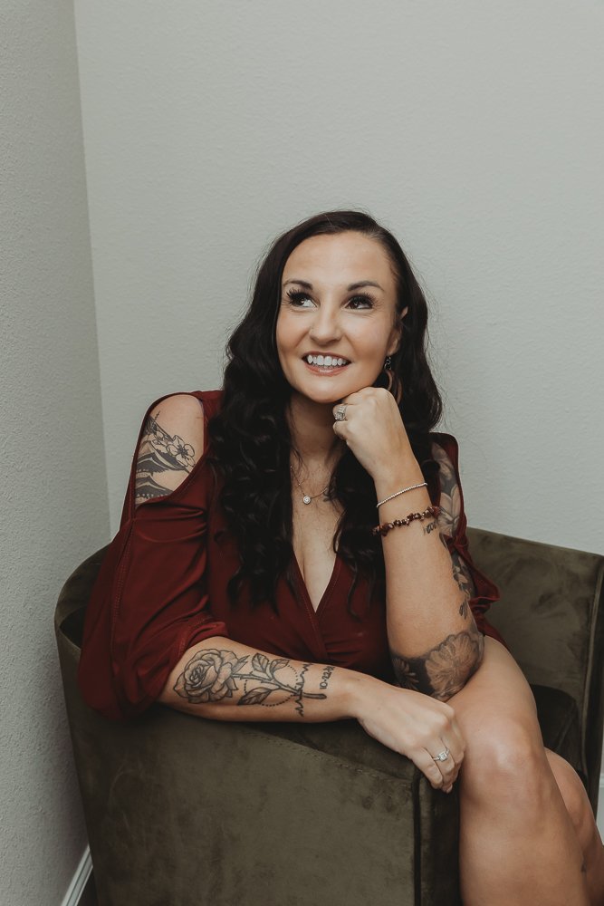 A woman with dark wavy hair sitting on a beige armchair, smiling, wearing a burgundy dress with cold shoulder sleeves and showcasing tattoos on her arms. She is resting her chin on her right hand, with various jewelry including a ring, bracelet, and necklace.
