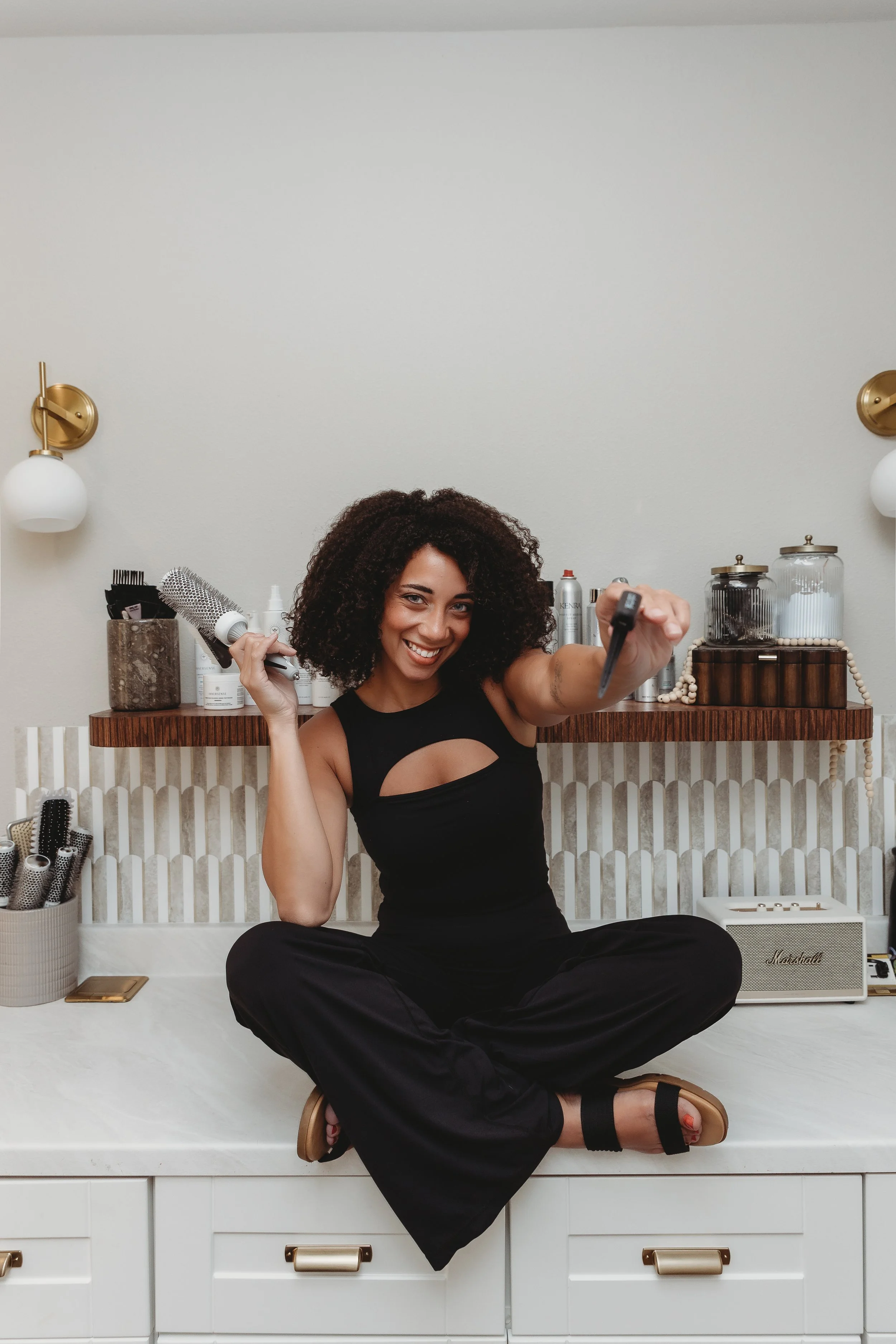 Woman sitting cross-legged on a white dresser, holding a hairbrush and a makeup spray bottle, smiling at the camera in a well-lit room with hair and beauty supplies on the shelves behind her.