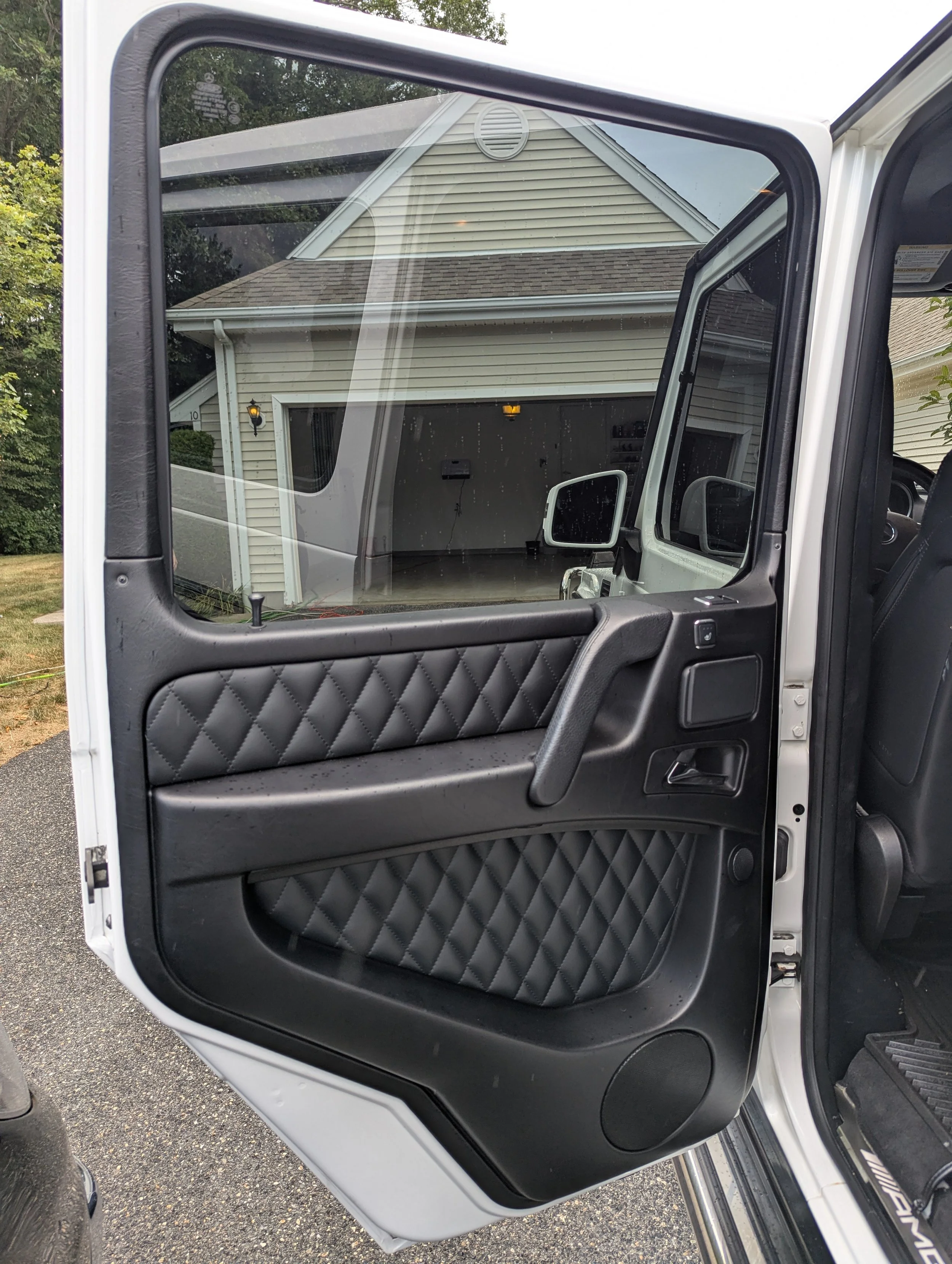 Close-up of an open vehicle door showing the inside panel with quilted black upholstery, door handle, window control, and speaker, with a house garage and driveway reflected in the window.