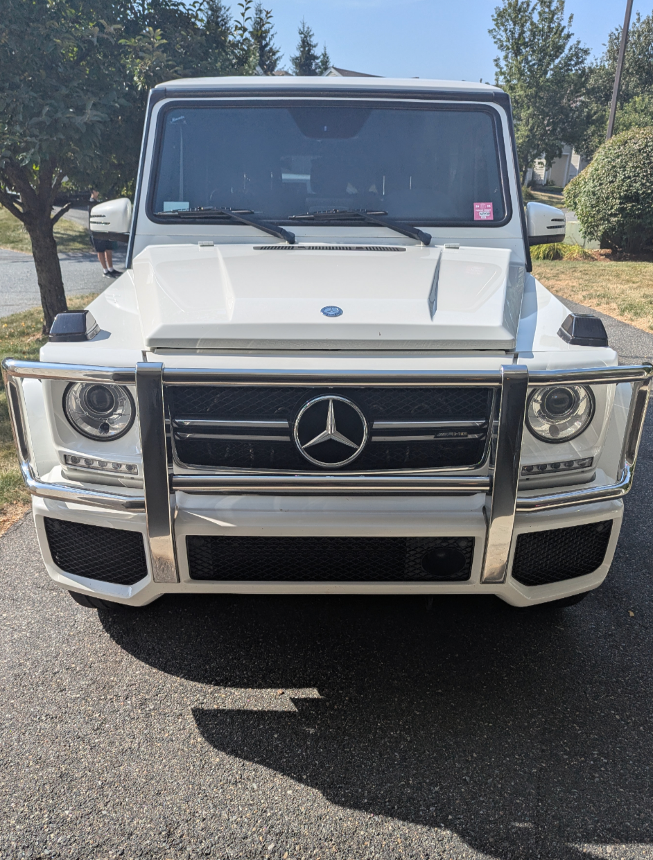 Front view of a white Mercedes-Benz G-Class vehicle parked on a driveway, with a grille featuring the Mercedes-Benz logo, round headlights, a chrome bumper guard, and a windshield with wipers.
