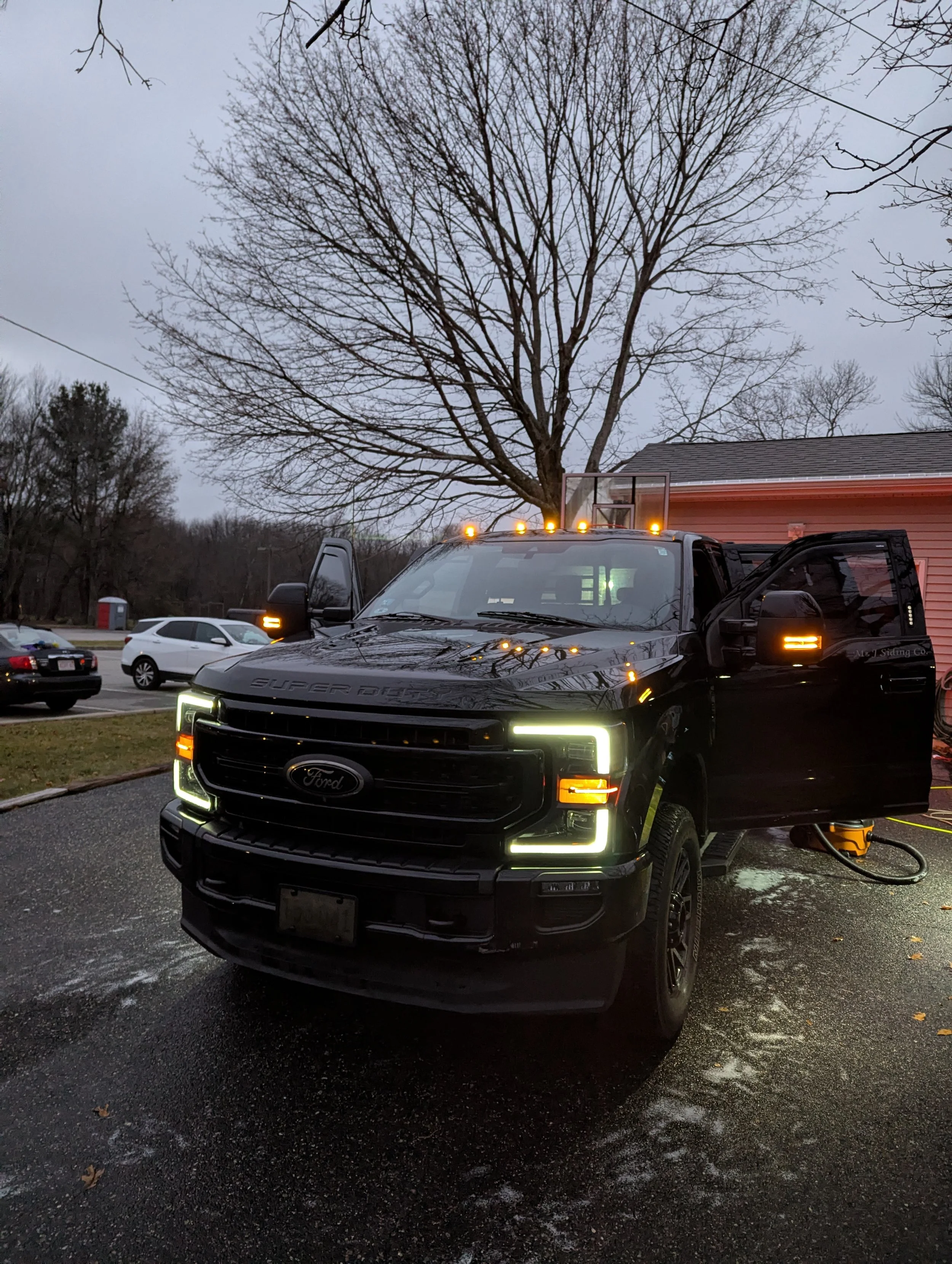 A black Ford Super Duty pickup truck with illuminated headlights and orange lights on top, parked outside near a pink house with one door open, and a large leafless tree in the background. The truck appears to be washing or detailing.