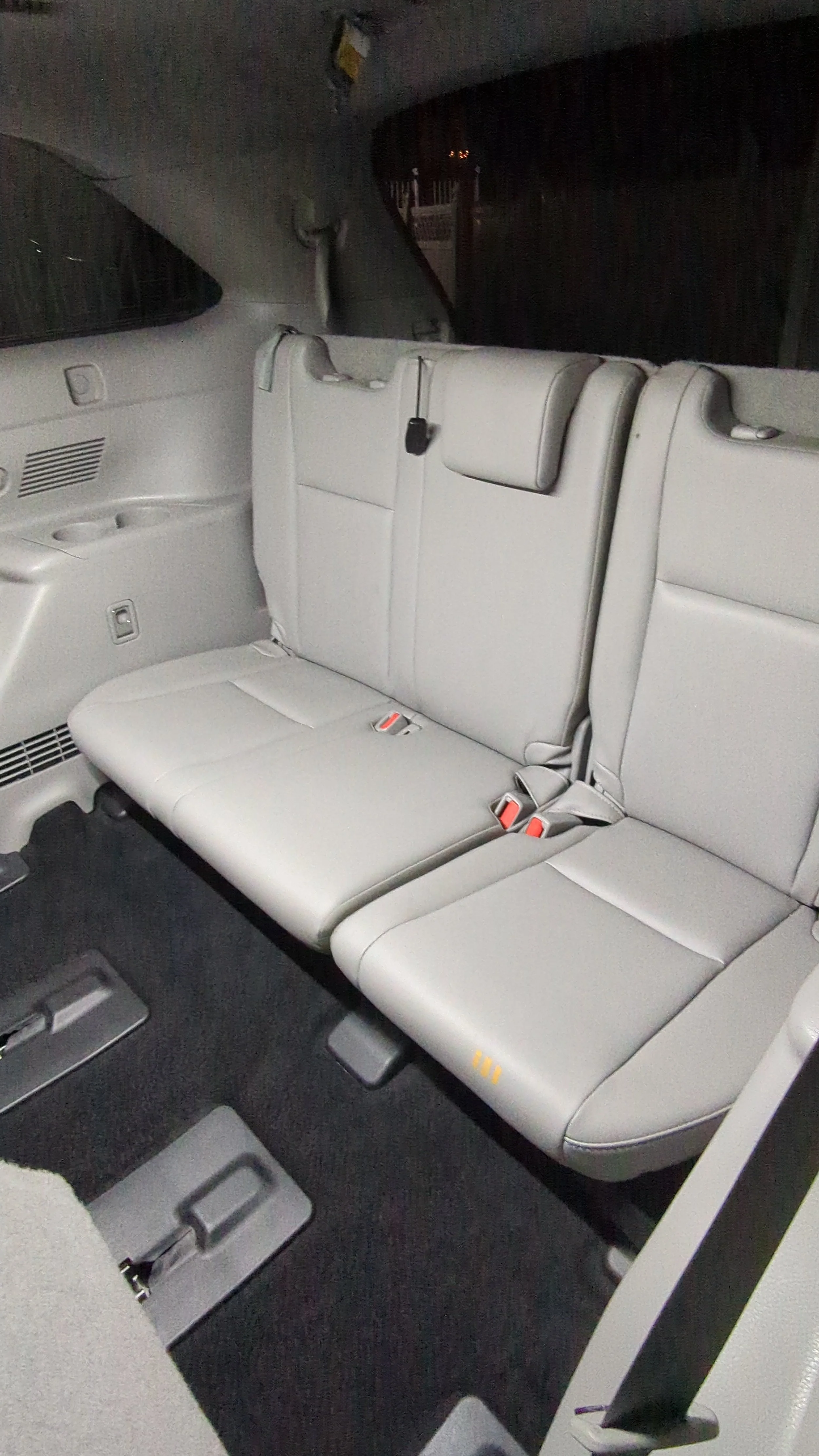 The back seats of a vehicle with grey upholstery, including a third-row bench seat with three headrests, seat belts, and a black interior wall.