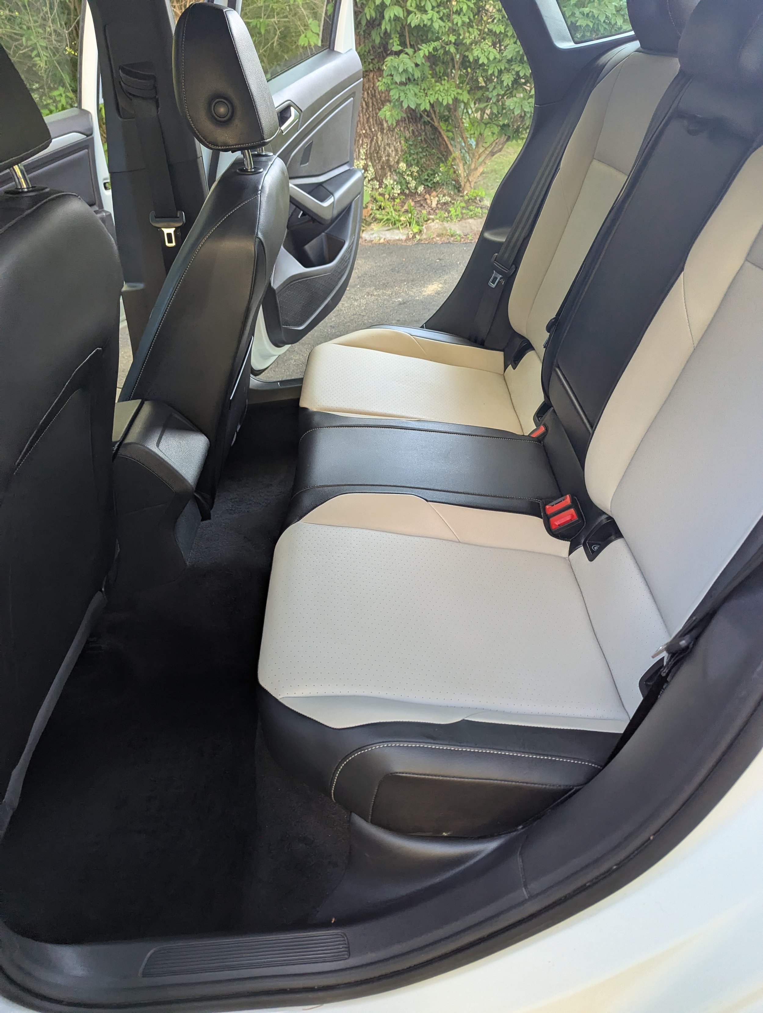 The back seat of a car with black and beige upholstery. The seats are clean and the environment outside shows greenery.