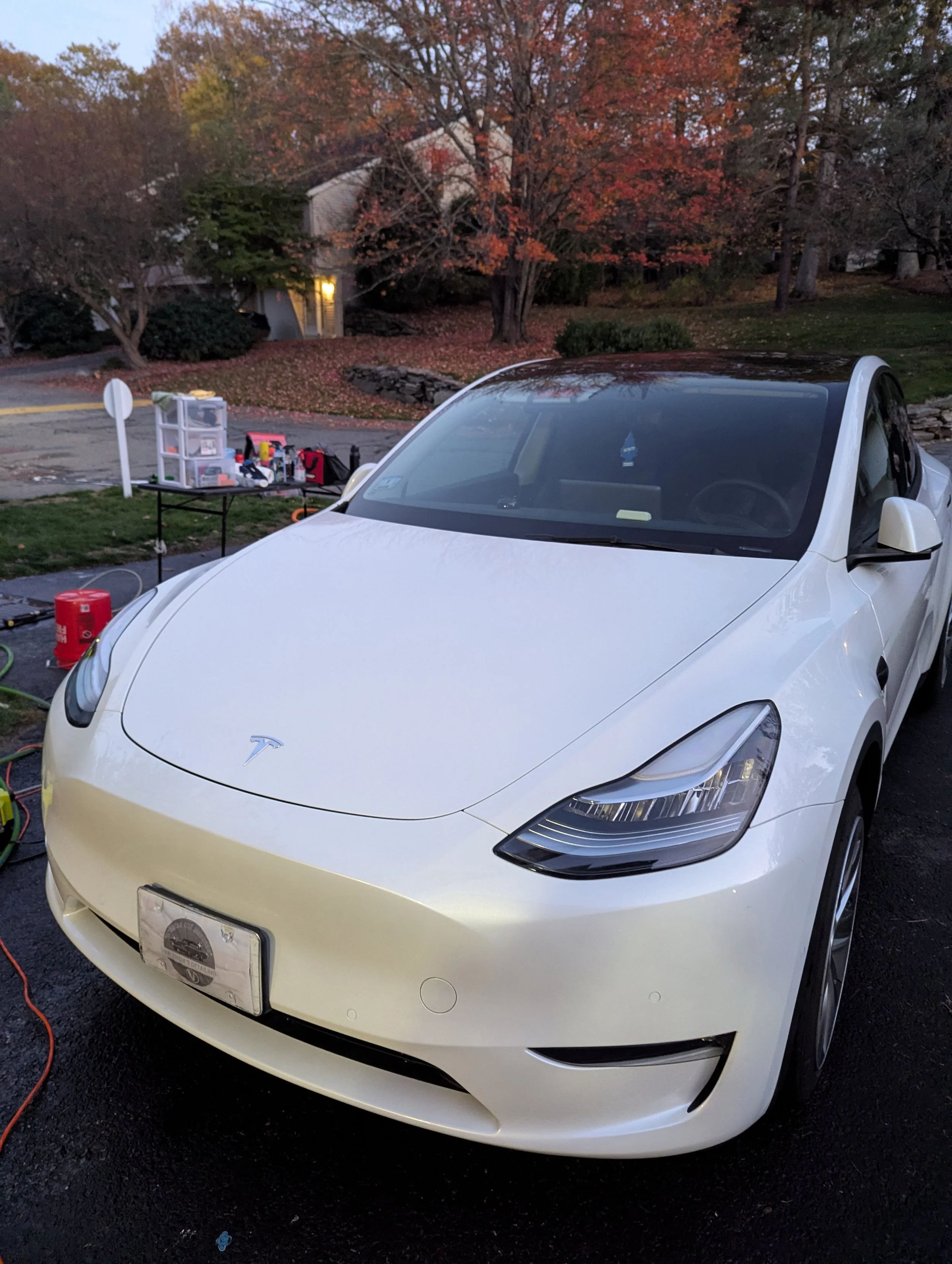 A white Tesla electric car parked outdoors with tools and equipment nearby, set against a backdrop of trees with fall foliage.
