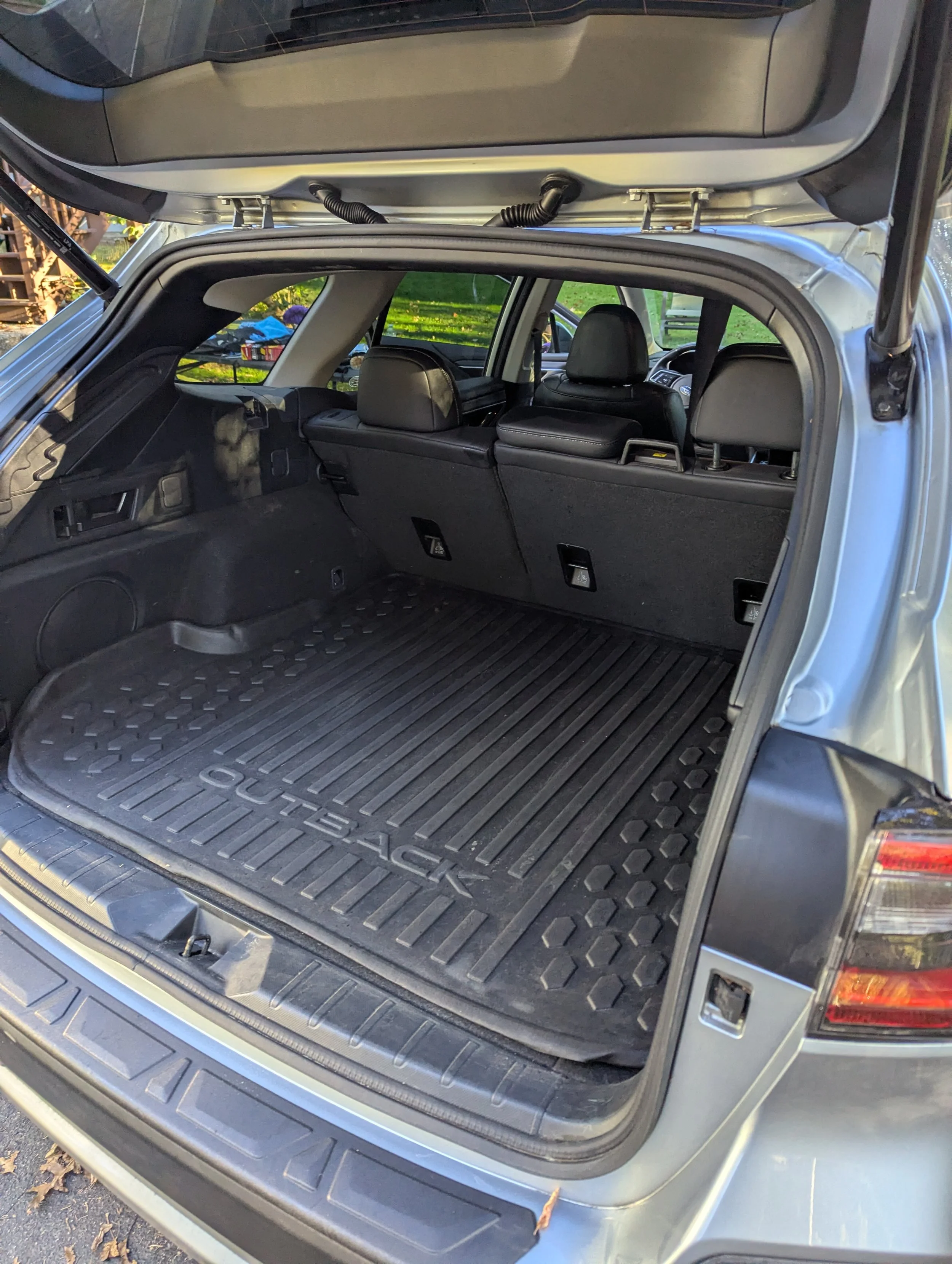 The open trunk of a gray Subaru Outback SUV, showing an empty cargo area with a black rubber mat, inside a residential outdoor setting.