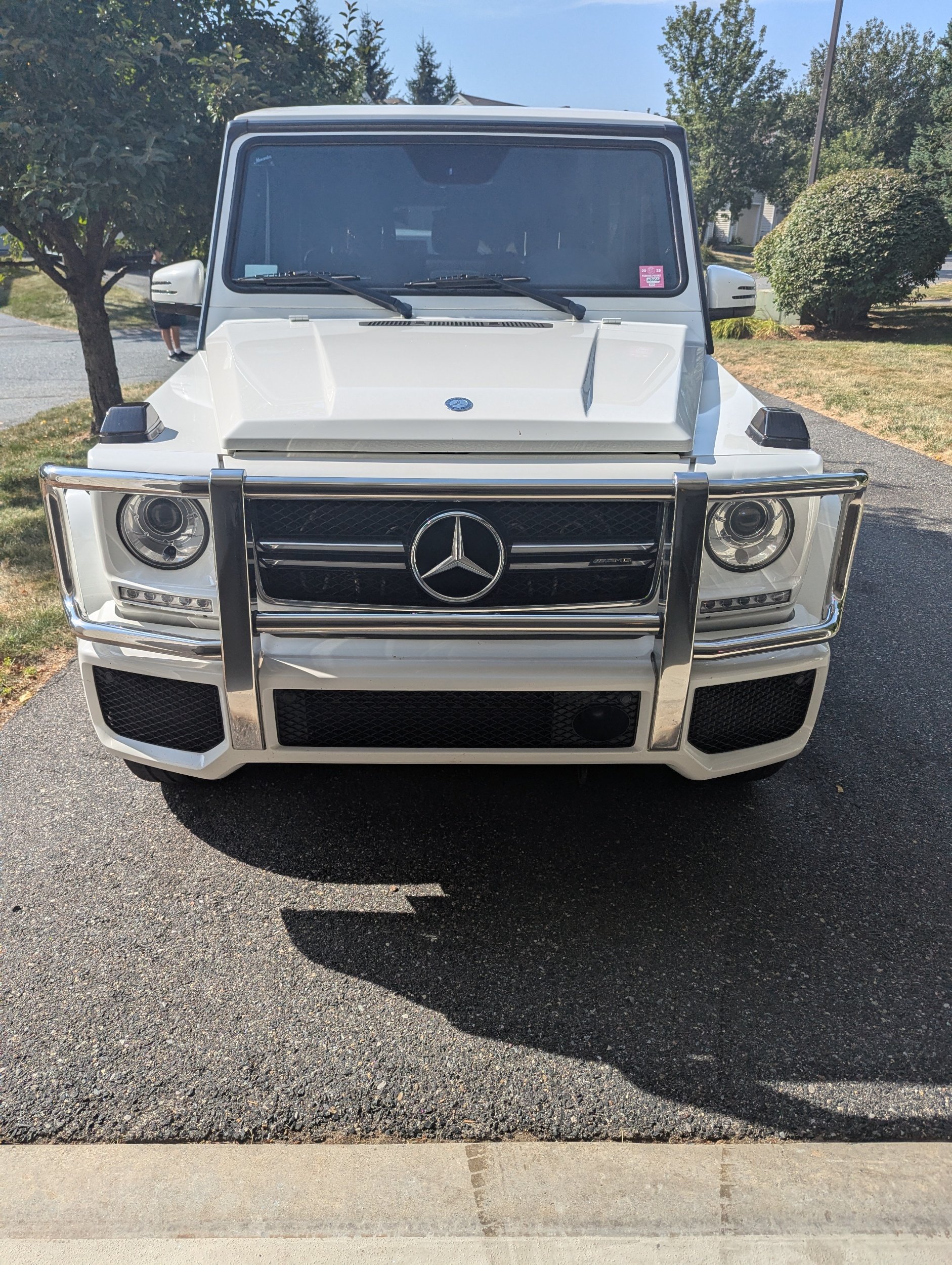 Front view of a white Mercedes-Benz G-Class SUV with a chrome bumper guard, parked on a driveway with trees and bushes in the background.