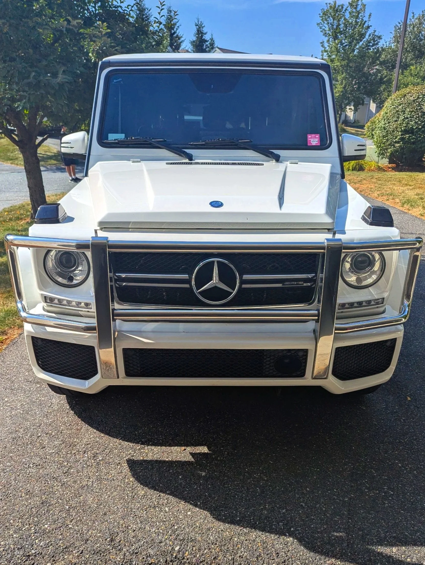 Front view of a white Mercedes-Benz G-Class SUV parked on a driveway, with trees and grass in the background.