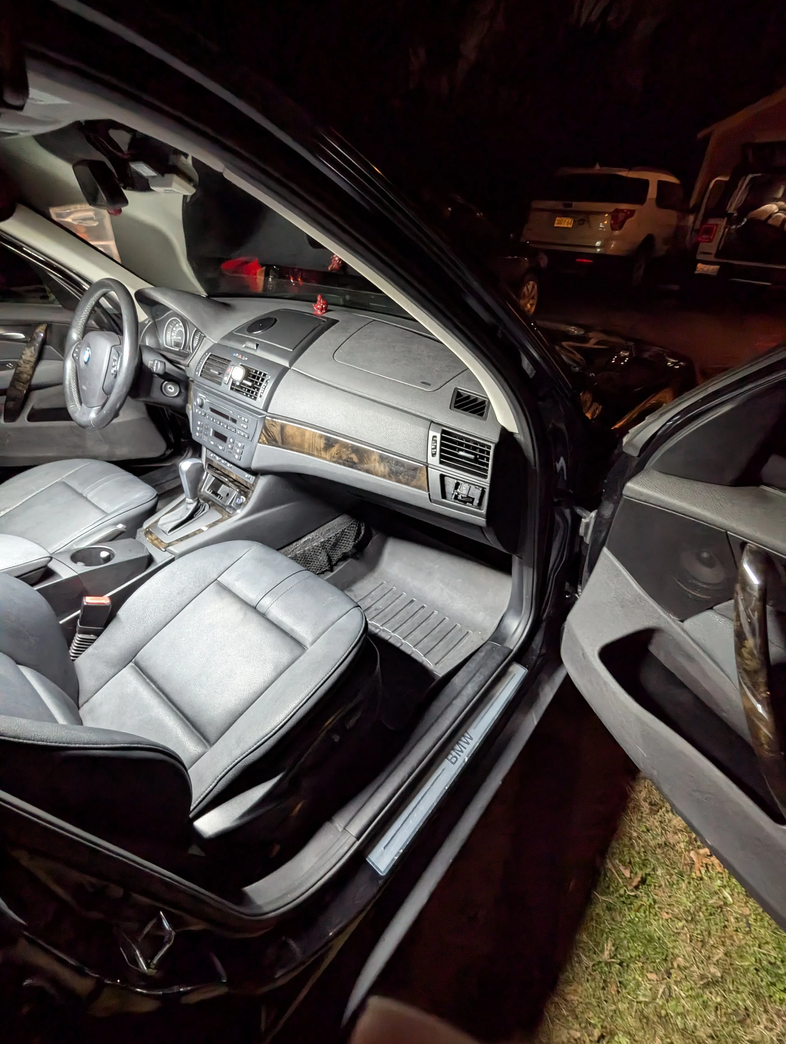 Interior of a black BMW vehicle, showing the front seats, dashboard, steering wheel, and open driver-side door at night, with other cars parked outside.
