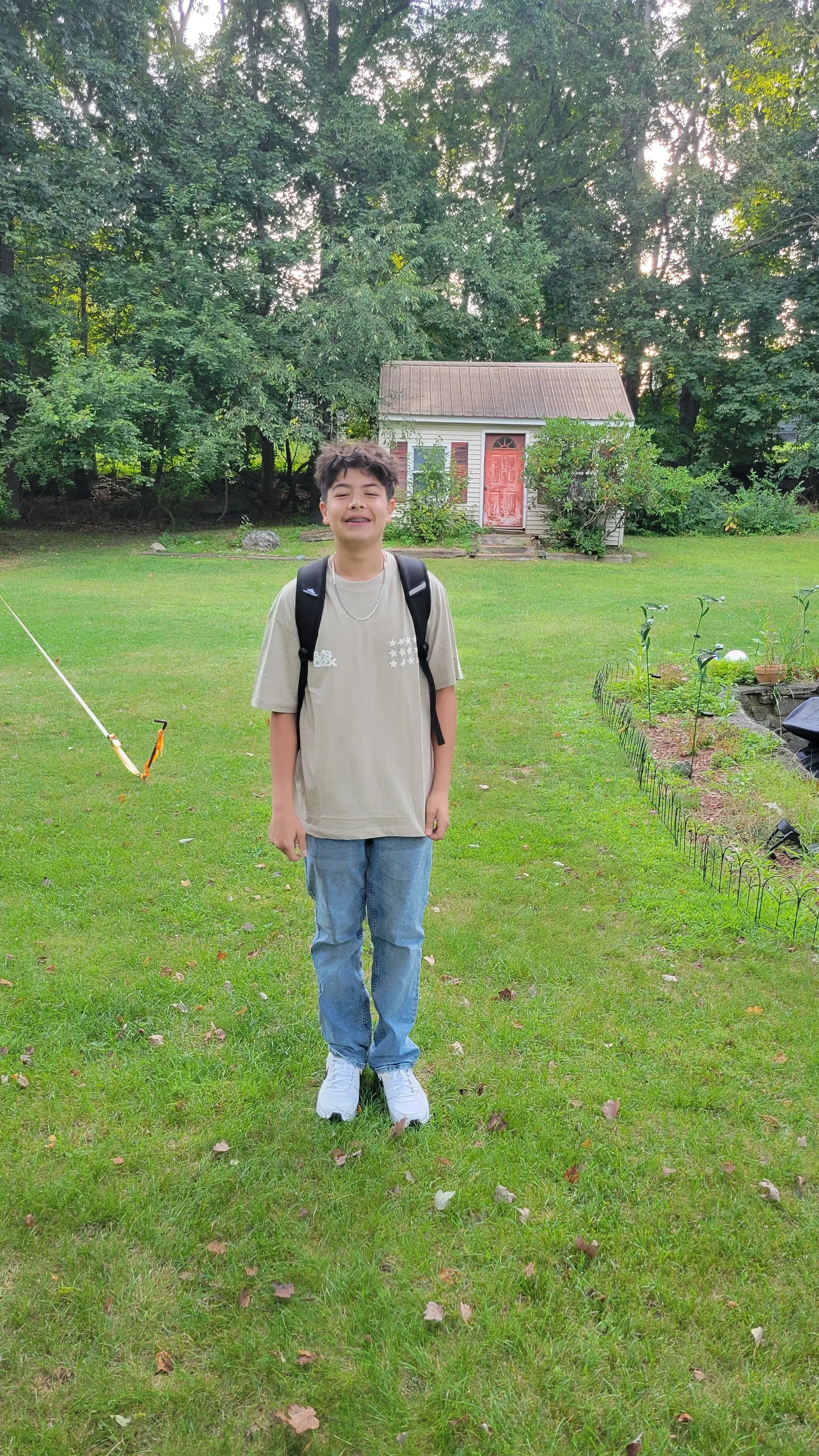 Young boy standing on green lawn with backpack, smiling, in front of small shed with red door, surrounded by trees and garden area.