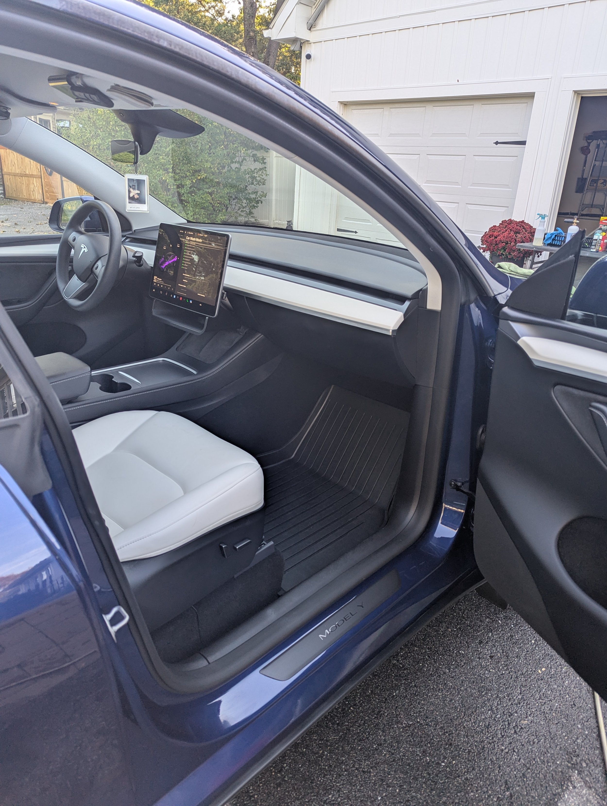 Interior of a blue Tesla Model Y, showing the driver seat with a white seat cover, a large central touchscreen display, and a small rearview mirror. The outside garage door and some gardening supplies are visible in the background.