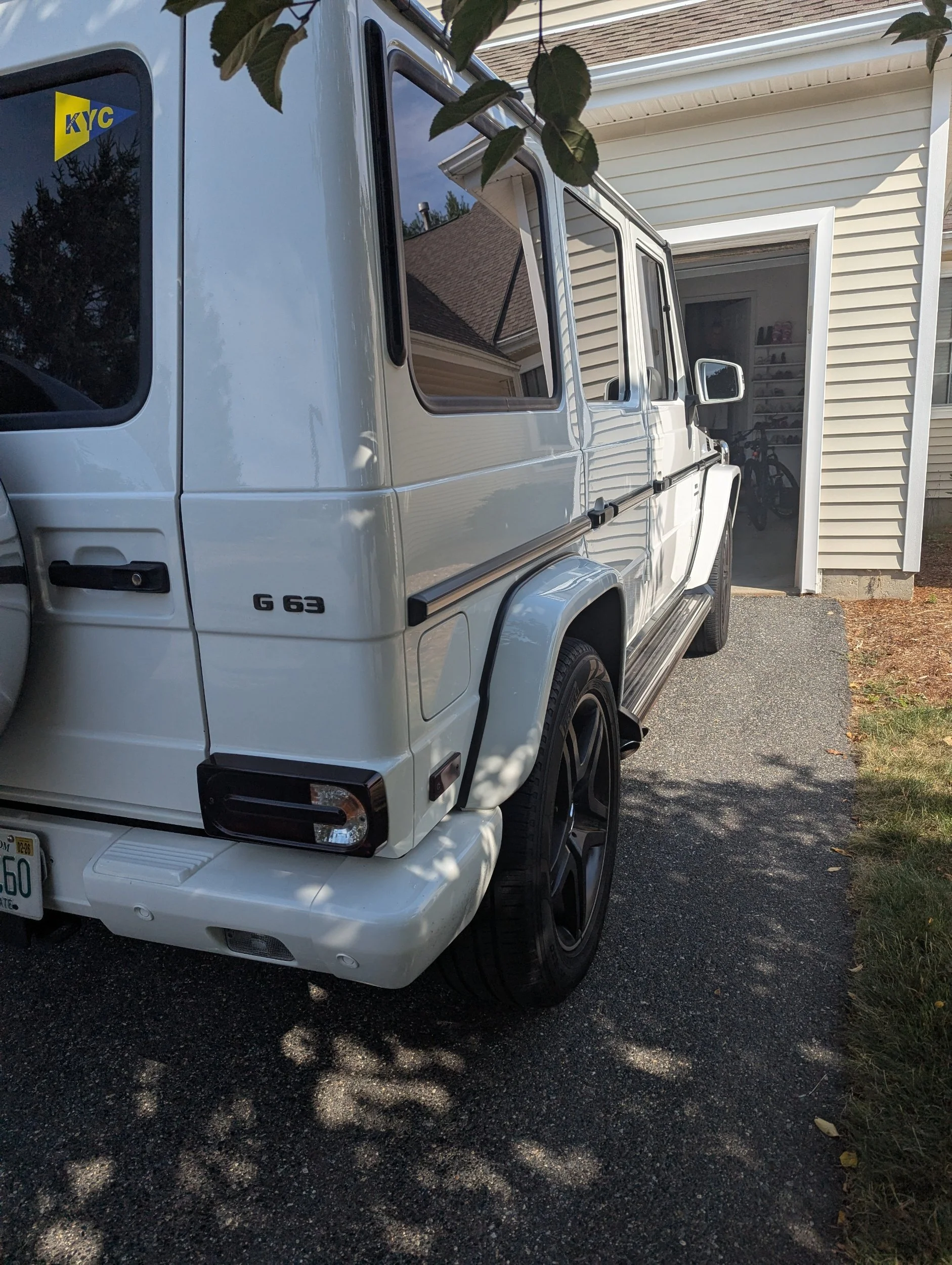 White Mercedes G63 parked on a driveway in front of a garage with bicycle and shoes inside. Tree branches above cast shadows on the vehicle.