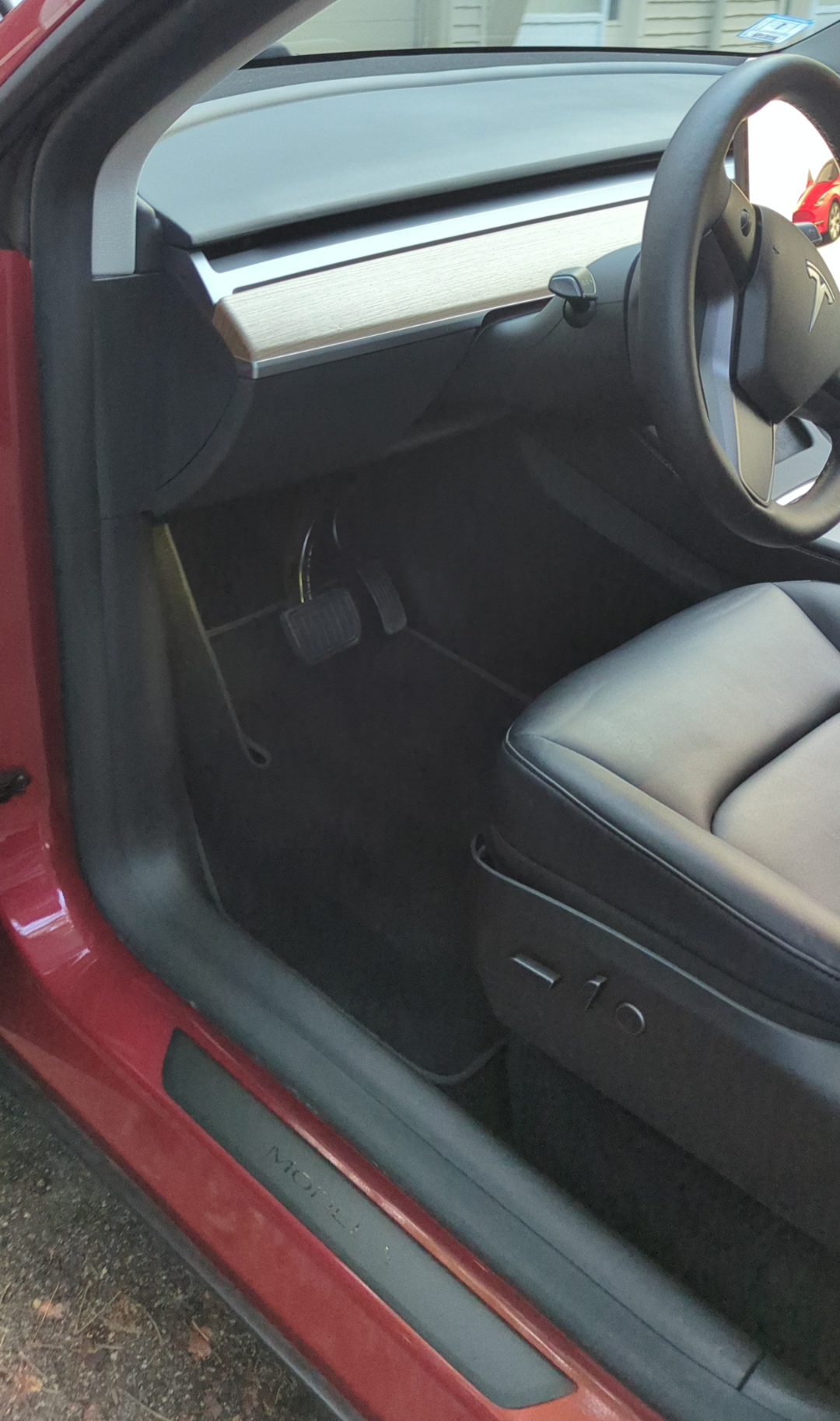 Interior of a Tesla Model 3 showing the driver's side, including the steering wheel with Tesla logo, pedals, and a minimalist dashboard with a wood trim.