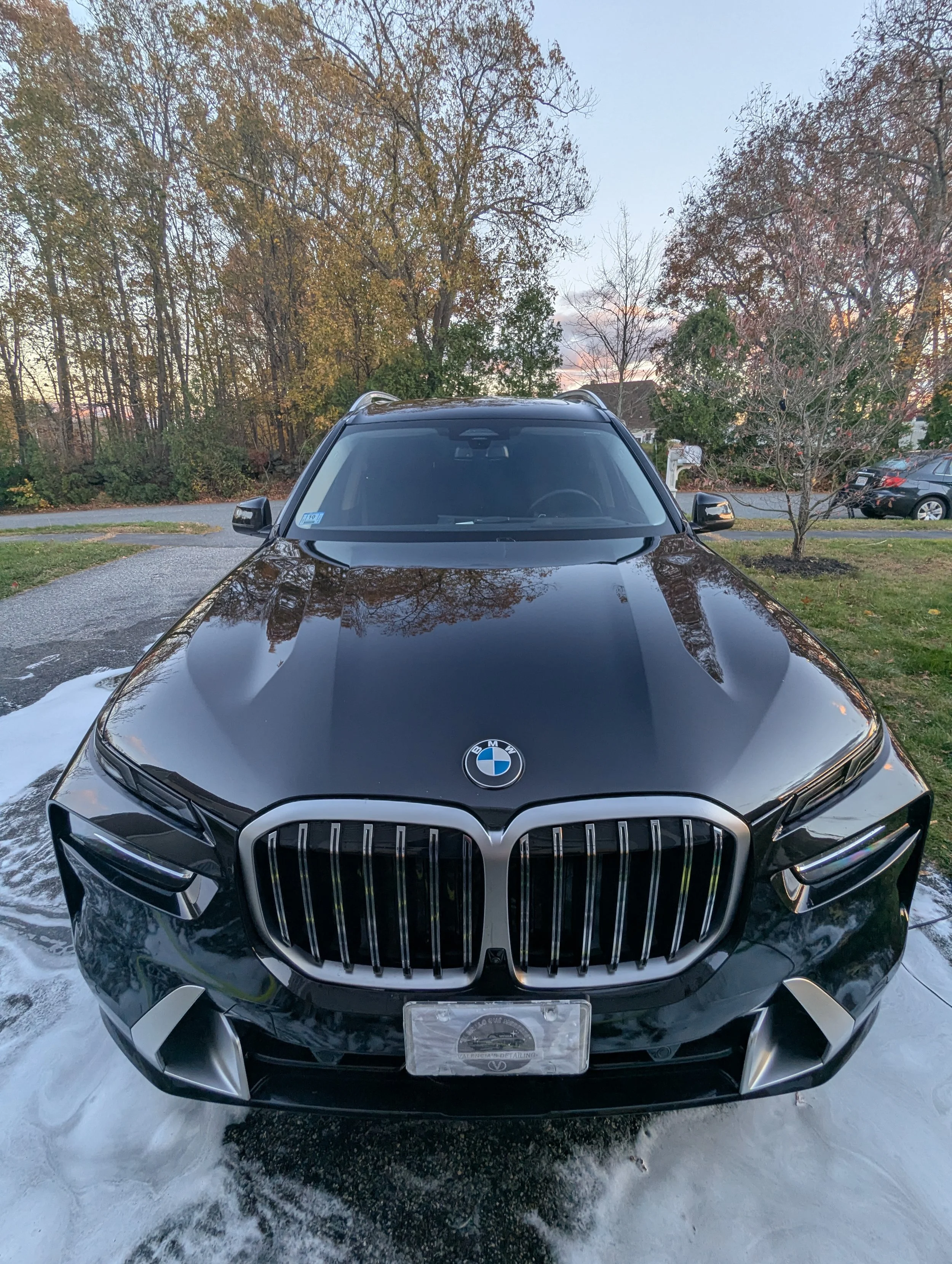 Black BMW SUV parked on a driveway with soap suds around the base, trees with fall foliage in the background, and a clear sky at sunset.