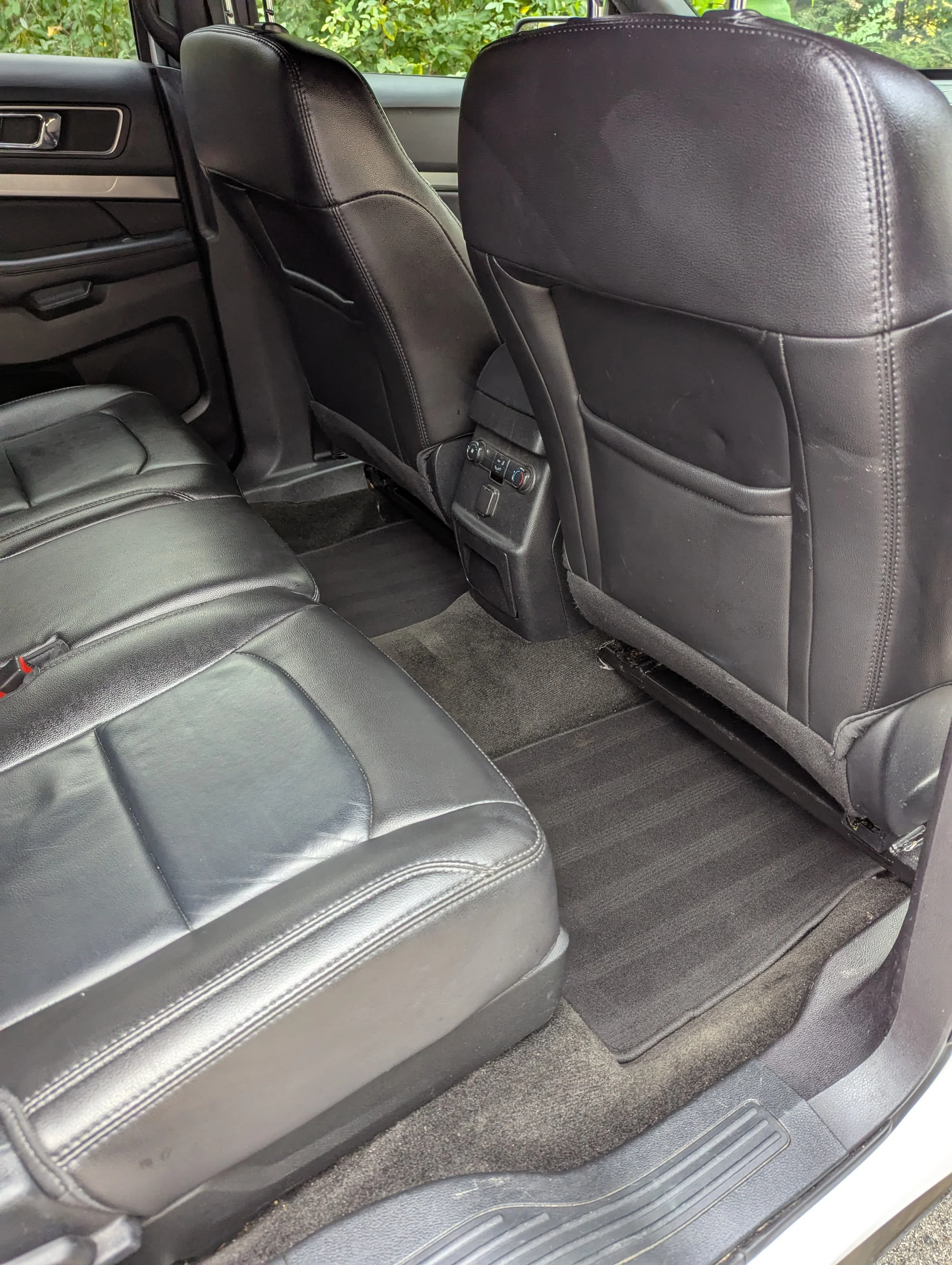 Interior of a vehicle showing the backseat and front seats with black leather upholstery, along with the back of the driver's and passenger's seats, floor mats, and some control buttons on the center console.