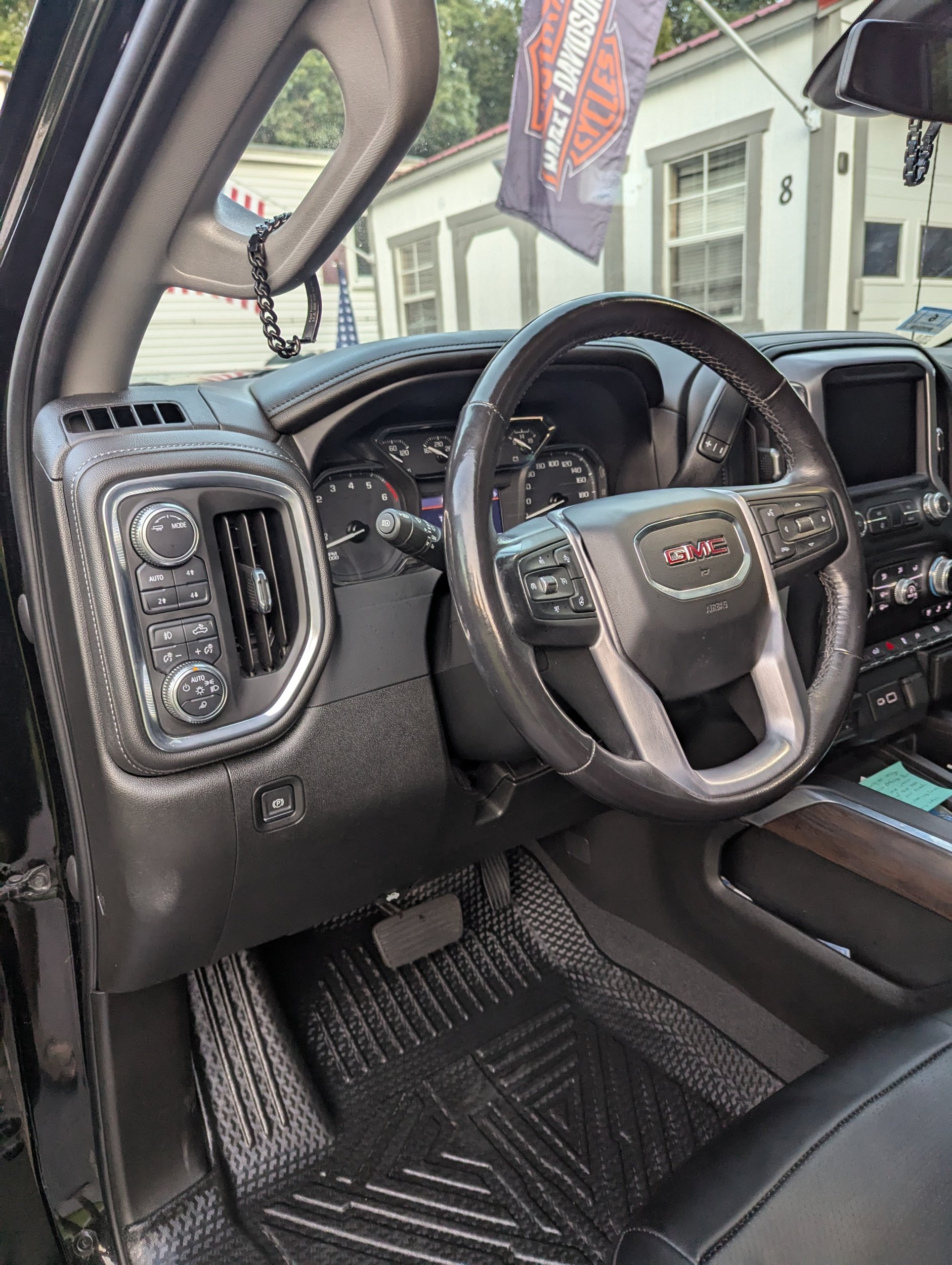 Inside view of a GMC vehicle dashboard, showing the steering wheel, dashboard gauges, control buttons, air vents, and a rearview mirror with a decorative chain hanging from it.