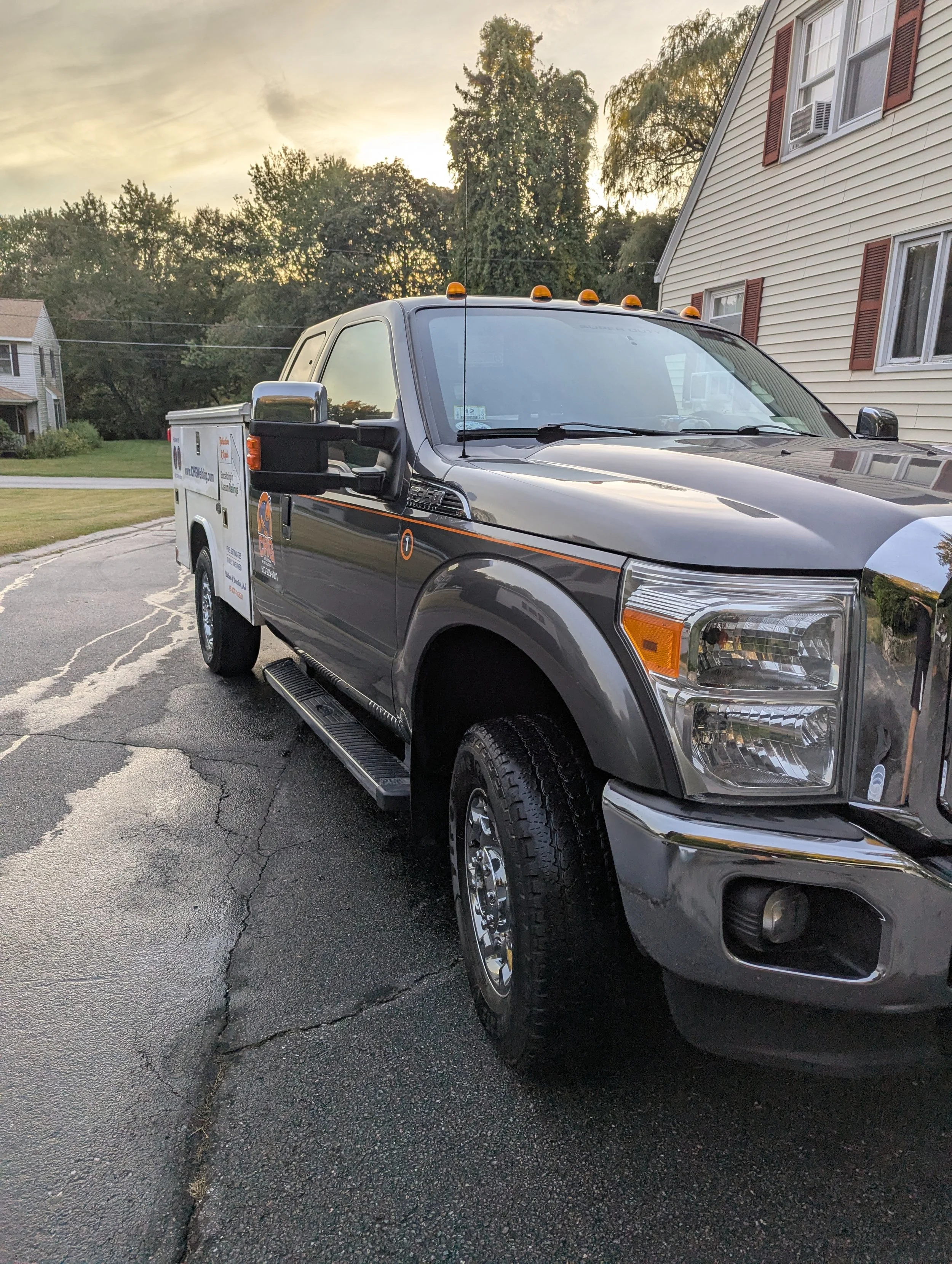 A black and gray service truck parked on a wet driveway with a house and trees in the background during sunset.