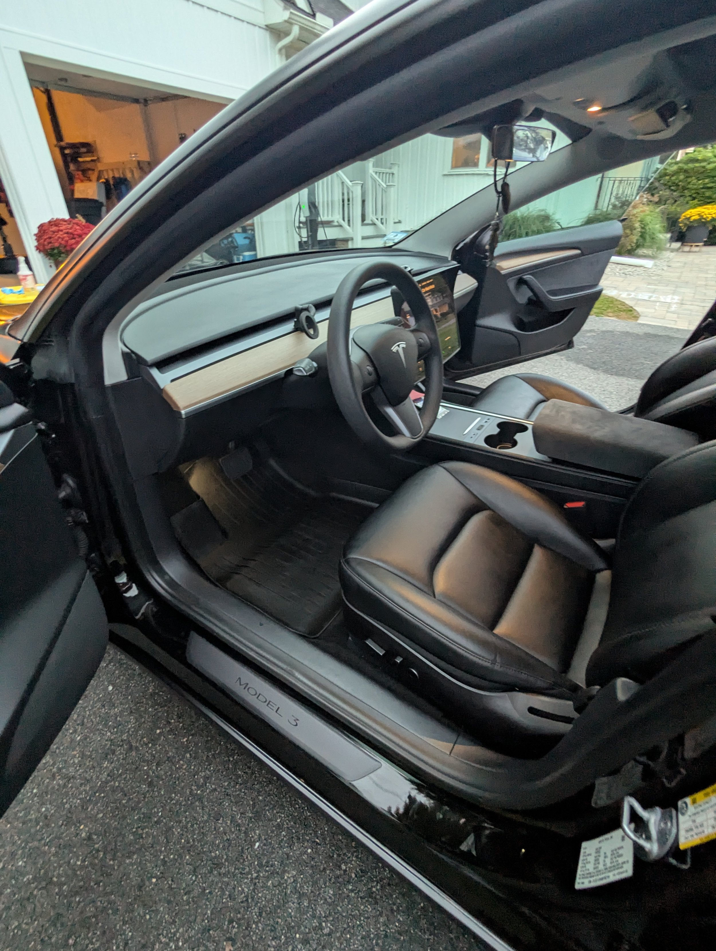Interior of a Tesla Model 3 electric vehicle, showing a black leather driver's seat, a dashboard with a central touchscreen display, and a steering wheel with the Tesla logo.