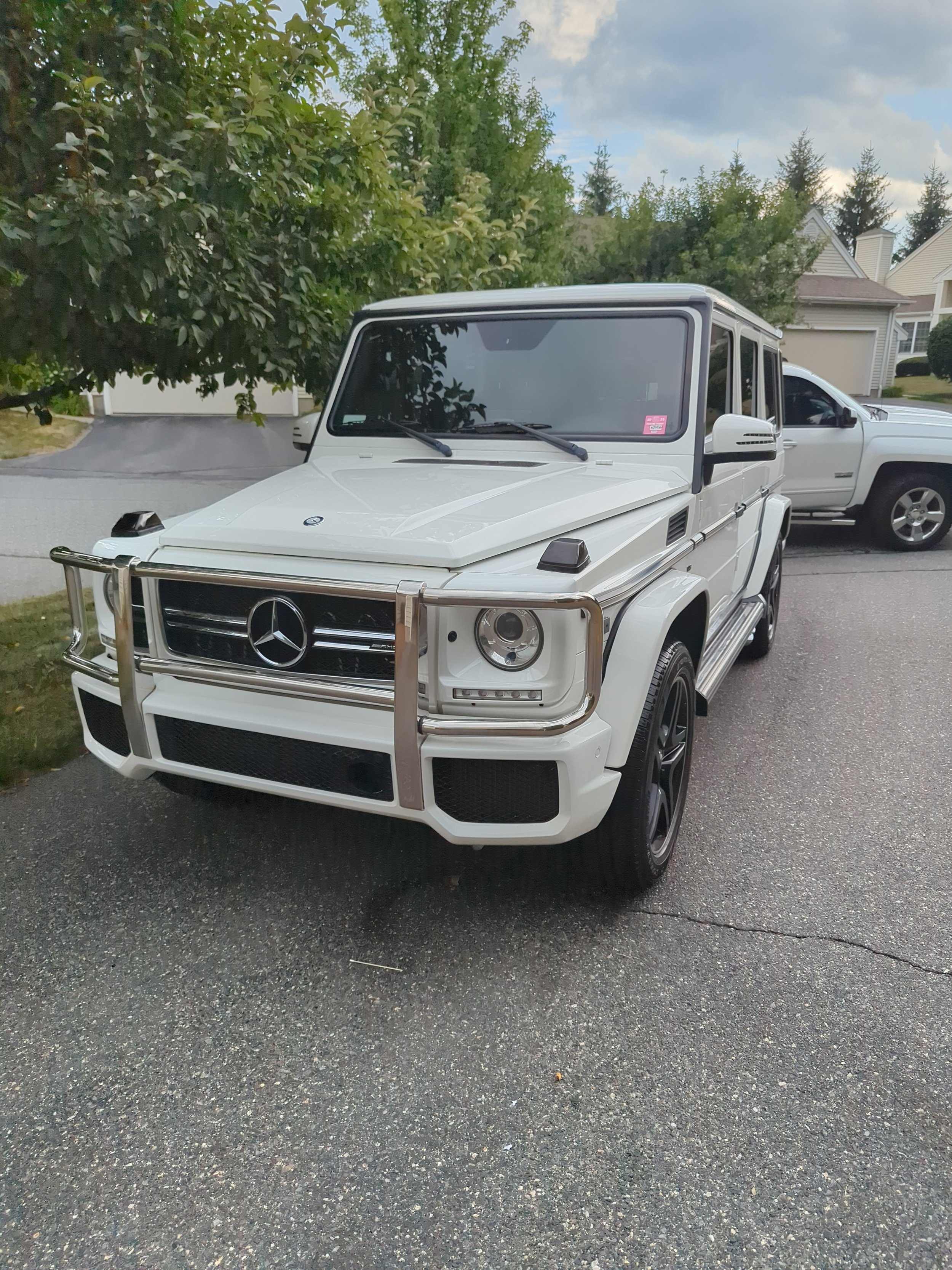 White Mercedes-Benz G-Class SUV parked on a driveway with trees and houses in the background.