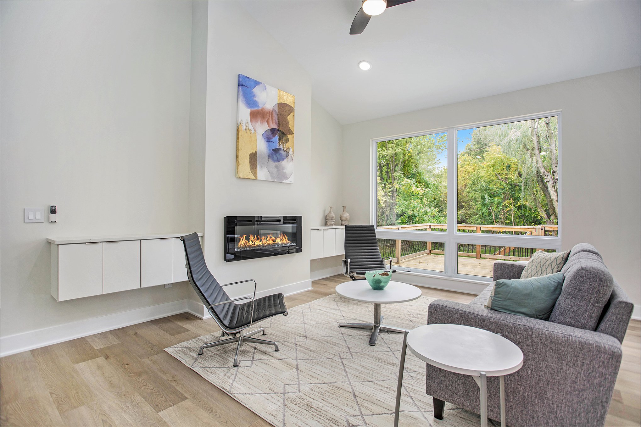 Living room with large windows showing outdoor greenery, gray sofa with cushions, black armchairs, a small white side table, a modern fireplace, and abstract wall art.