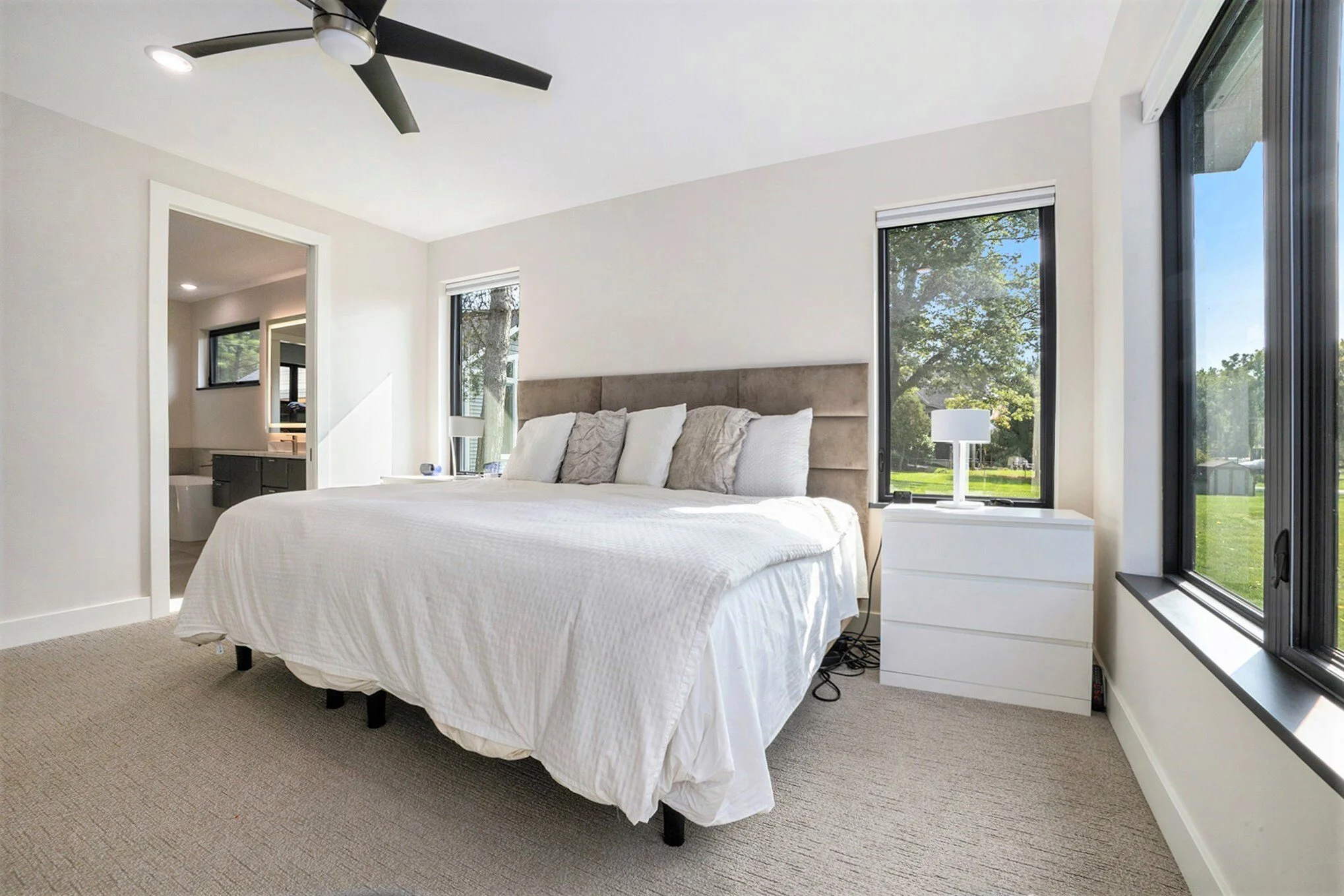 A bedroom with a large bed, beige headboard, white bedding, and several pillows. There are three windows with black frames and white blinds, a white nightstand with a lamp, and an open doorway leading to a bathroom. The room has beige carpet, white w