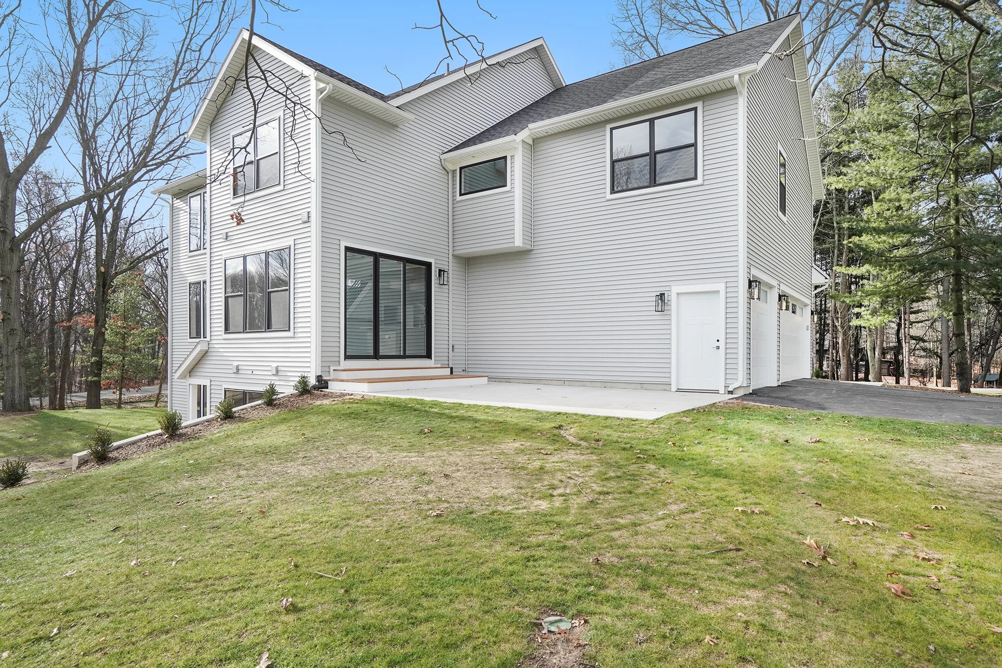 A modern two-story house with white siding, black window frames, a gray roof, and a concrete patio at the back. There are trees surrounding the house and a green lawn in the foreground.