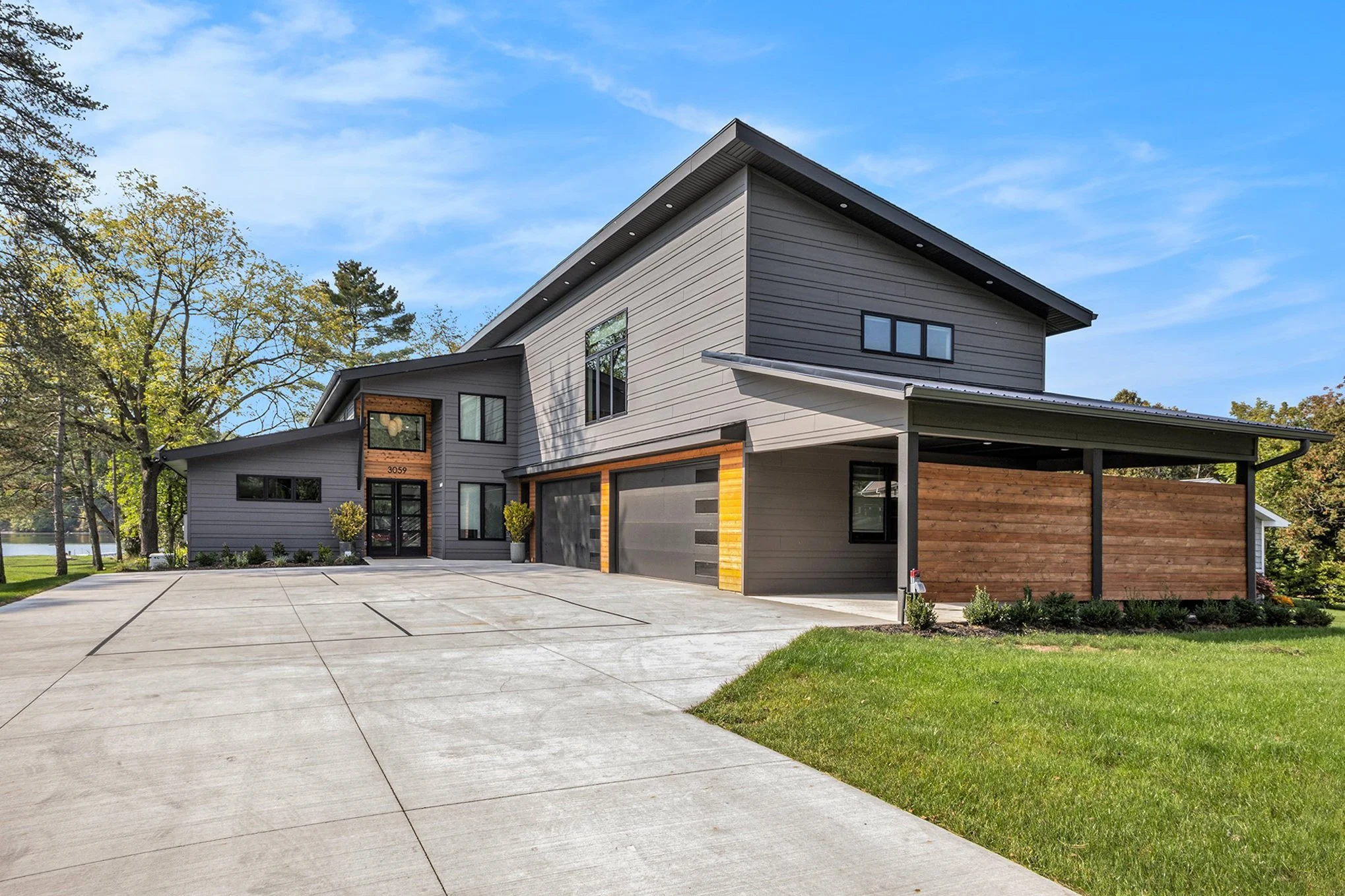 Modern two-story house with gray siding, black accents, and a large driveway, surrounded by trees and green lawn, under a blue sky with some clouds.
