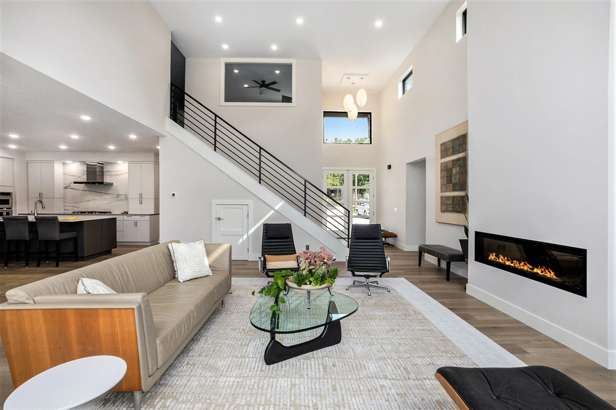 Modern open-concept living room with beige sofa, black lounge chairs, glass coffee table with plant, white fireplace, and kitchen in background.