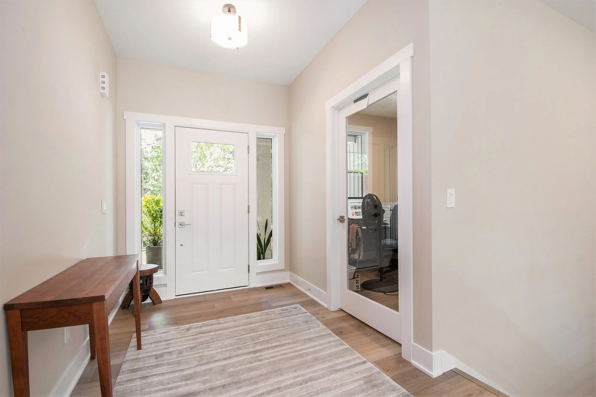 Entryway with white front door, glass side windows, a side door with glass, a wooden console table, a small round stool, a beige rug, and an office visible through an open door.