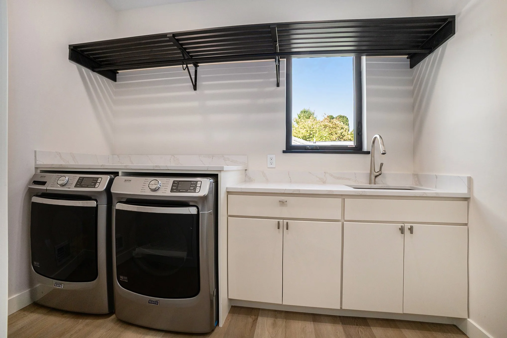 Laundry room with a front-loading washing machine and dryer, a white countertop with a sink, a window with a view of trees, and a black wall-mounted drying rack.