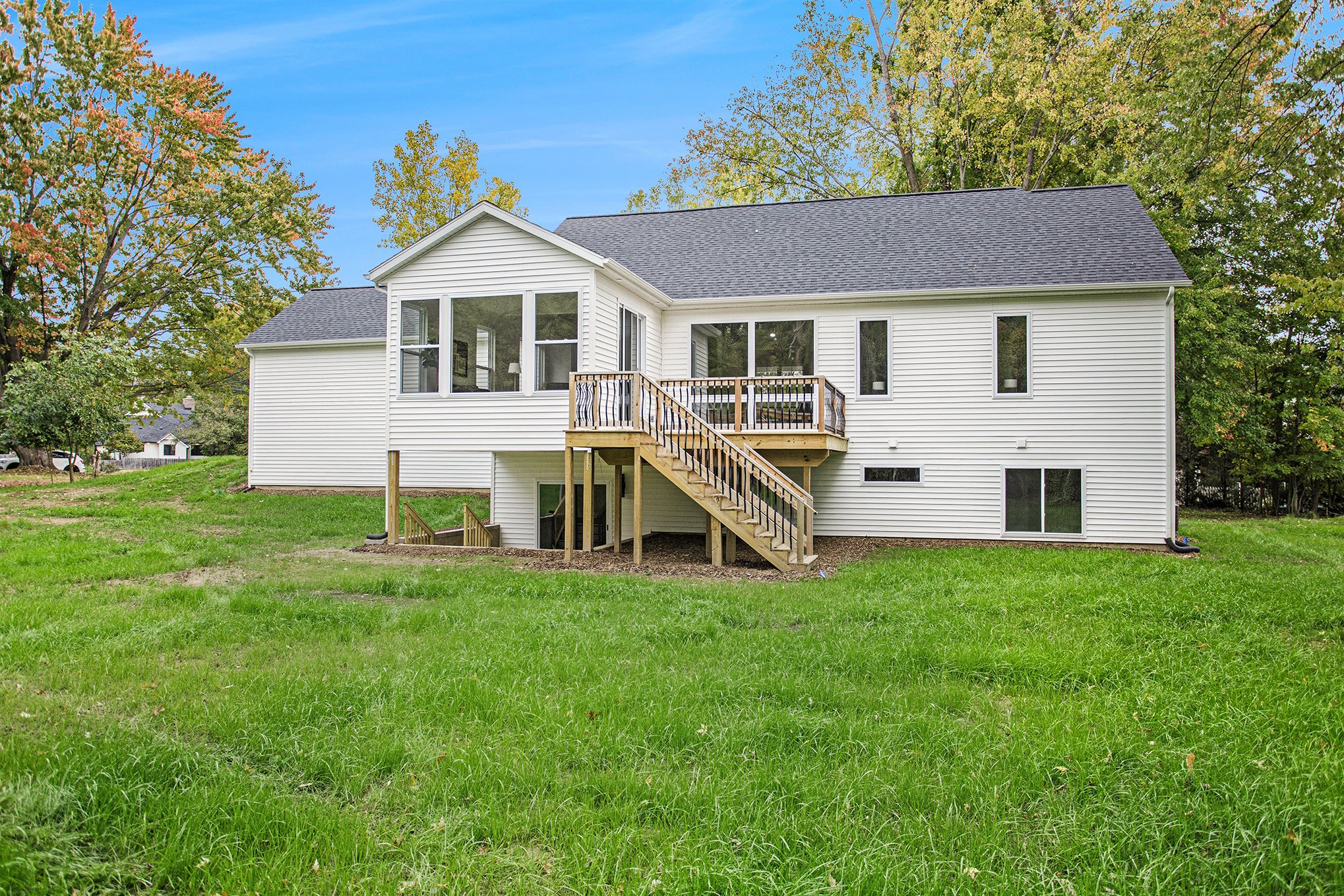 Back of a white house with a deck and stairs leading down to a grassy yard, surrounded by trees with autumn foliage, under a clear blue sky.