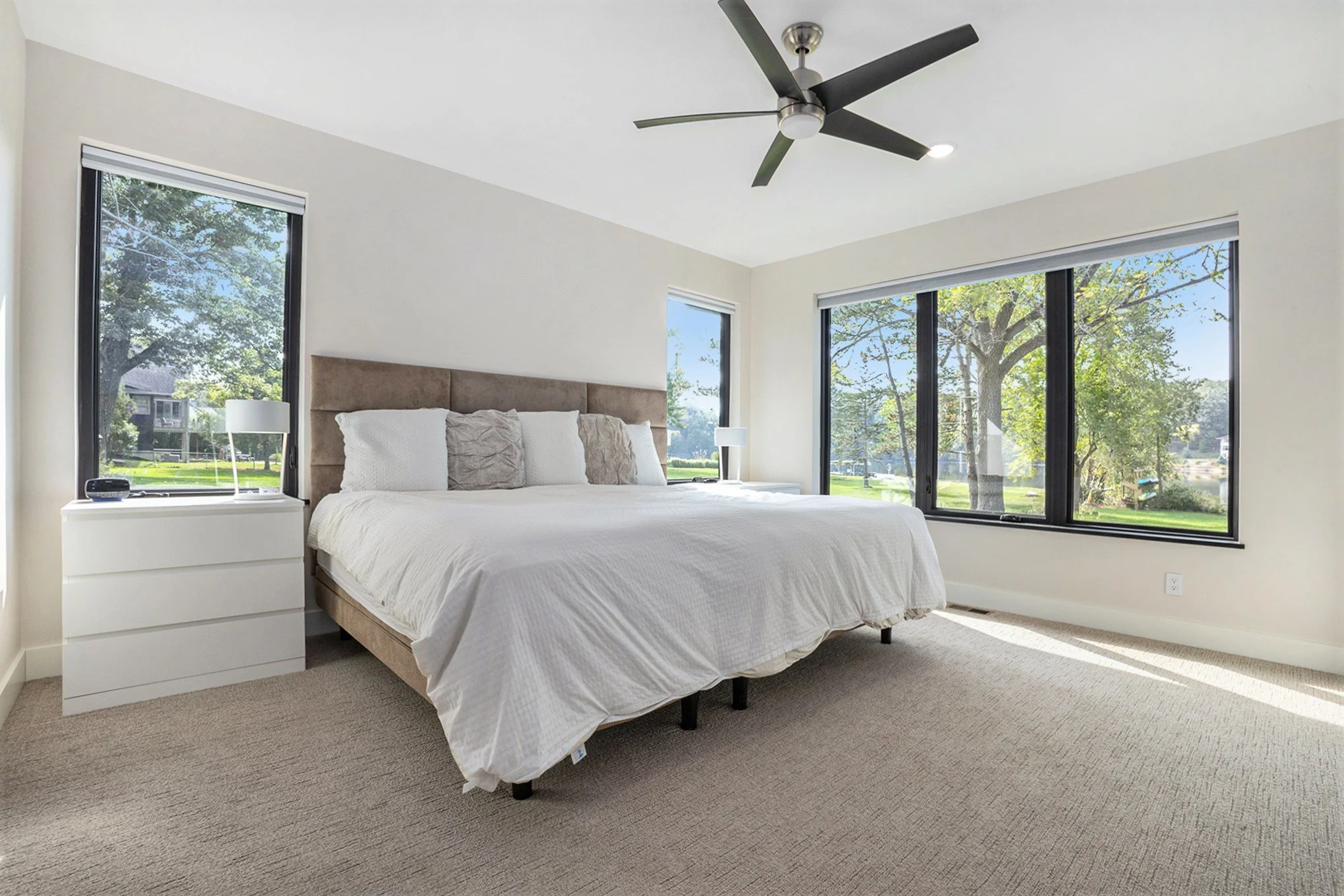 Bright bedroom with large windows showing greenery outside, a white bed with a beige headboard, white bedding, and two nightstands with lamps.