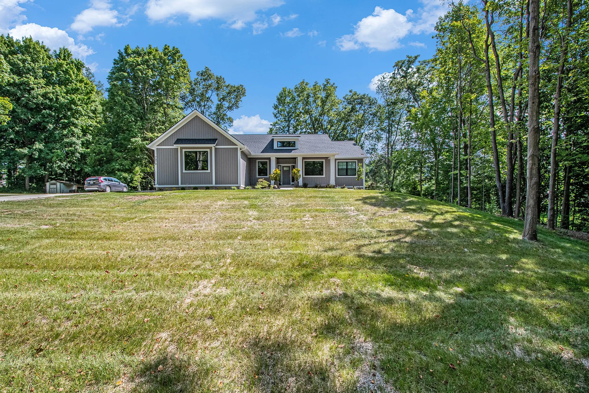 A modern gray house with large windows and a front door, situated on a hill with a grassy lawn and surrounded by tall trees under a blue sky with scattered clouds.