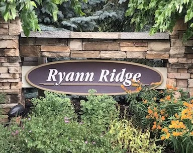 A sign reading 'Ryann Ridge' mounted on a stone wall surrounded by green plants and orange flowers.