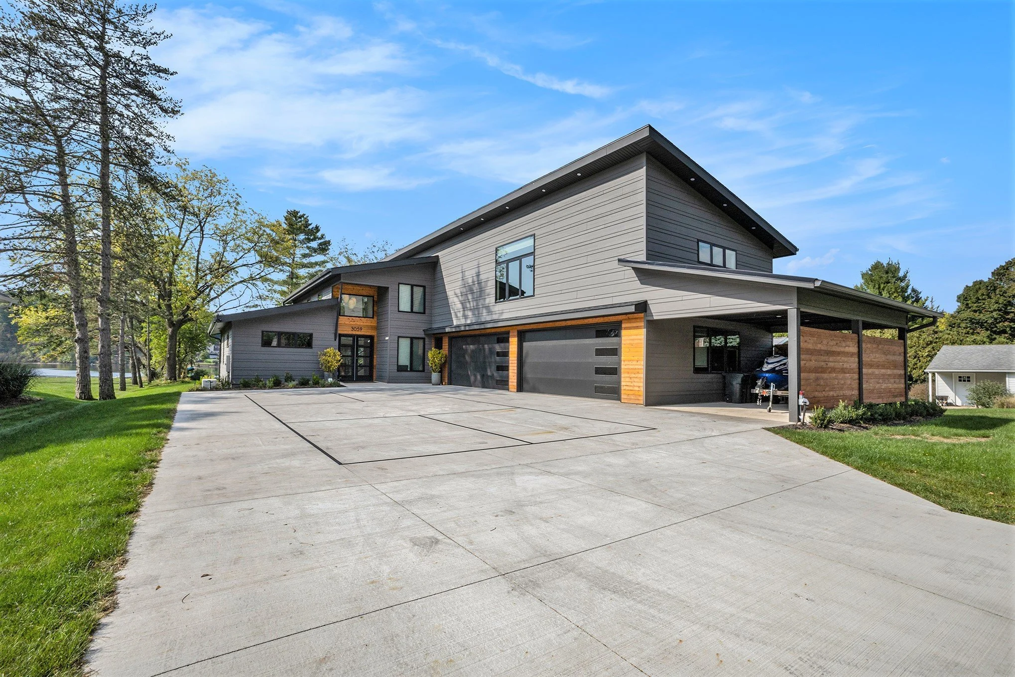 Modern two-story house with gray siding, black garage doors, and wooden accents, surrounded by a large concrete driveway, green lawn, and trees under a blue sky.