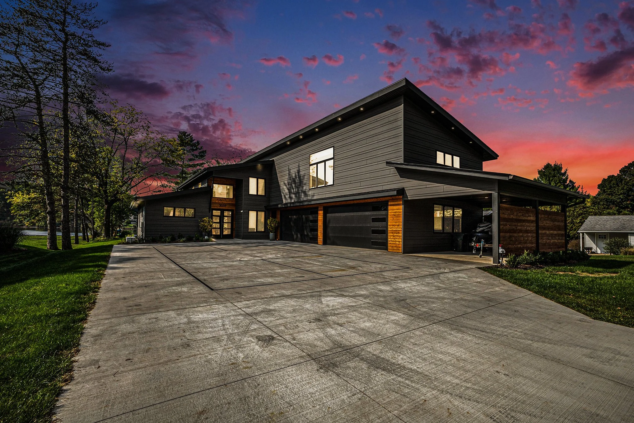 Modern two-story house with black exterior, wooden accents, and a garage, at sunset with a colorful sky and trees in the background.