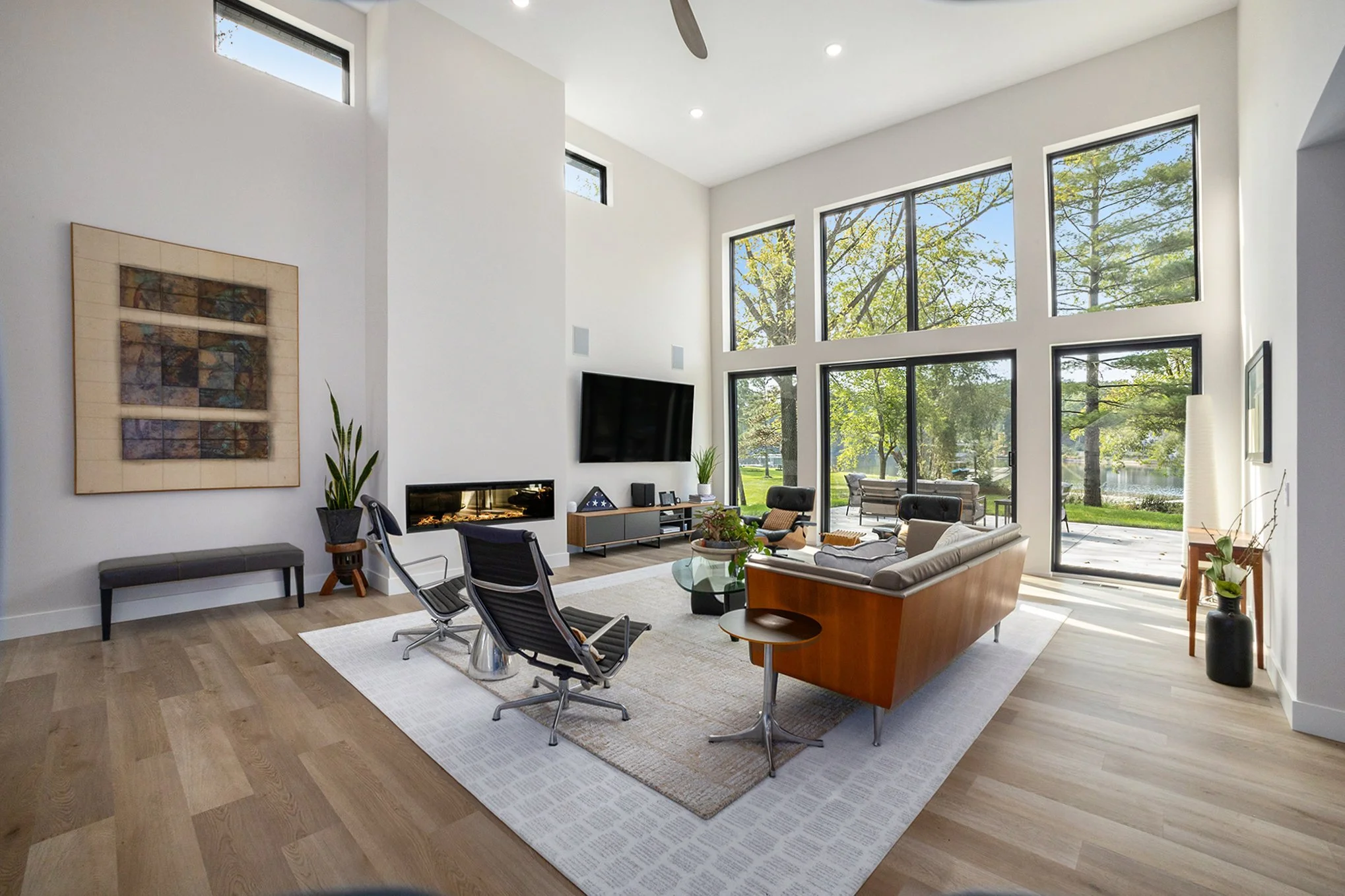 Modern living room with large windows, high ceiling, wooden flooring, and minimalistic decor, including a TV, fireplace, coffee table, and seating area.