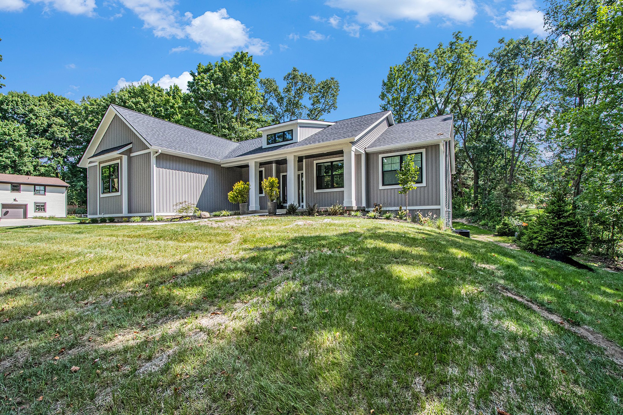 Modern single-family house with gray siding and a gabled roof, surrounded by a green lawn with trees and shrubs, under a blue sky with some clouds.
