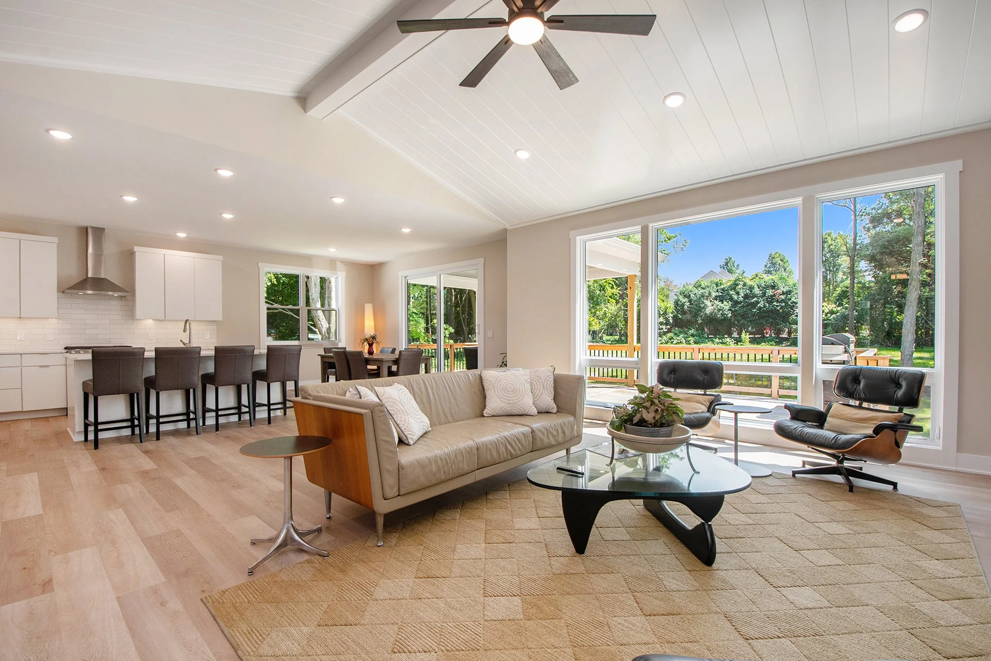 Bright living room with large windows showing greenery outside, featuring a beige sofa with white pillows, a glass coffee table, black lounge chairs, and a ceiling fan.