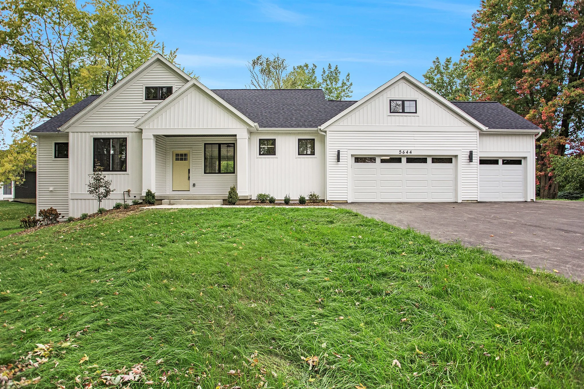 White modern house with a three-car garage, steps leading to a front door, surrounded by a green lawn and trees, under a partly cloudy sky.