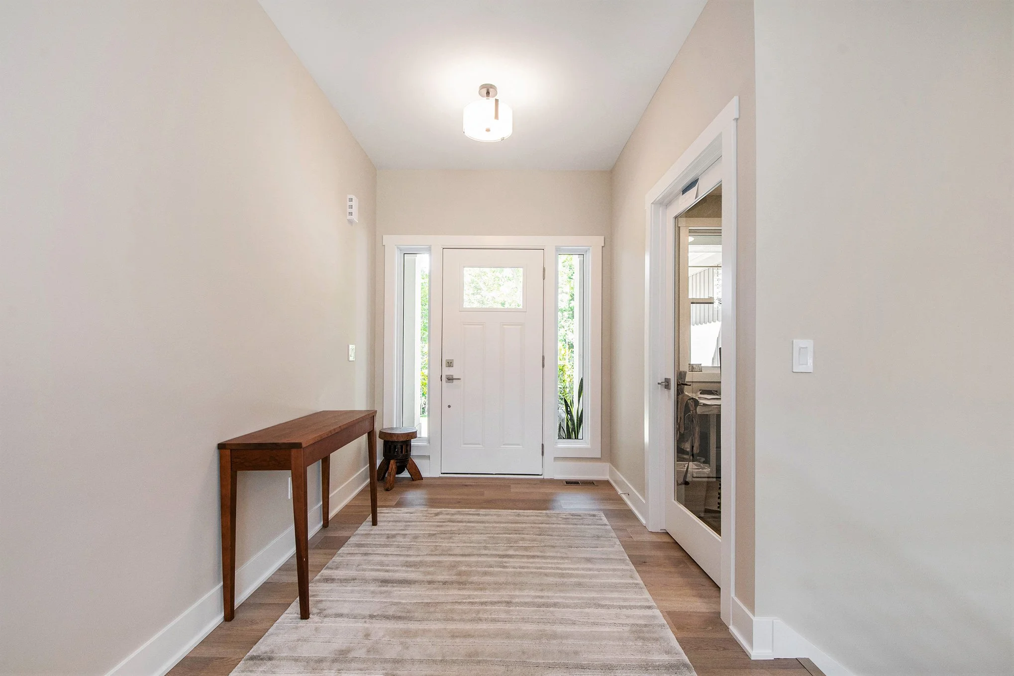 Entryway with front door, side windows, wooden console table, small stool, and beige area rug on hardwood floor.