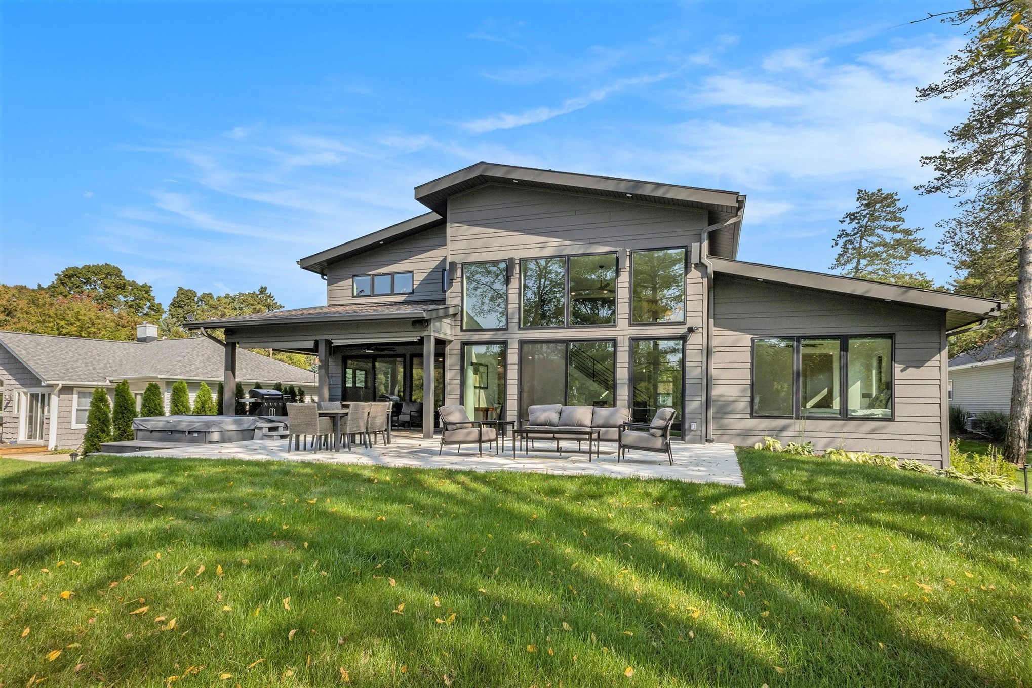 Modern two-story house with large glass windows and a spacious backyard patio with outdoor furniture, surrounding trees, and a clear blue sky.