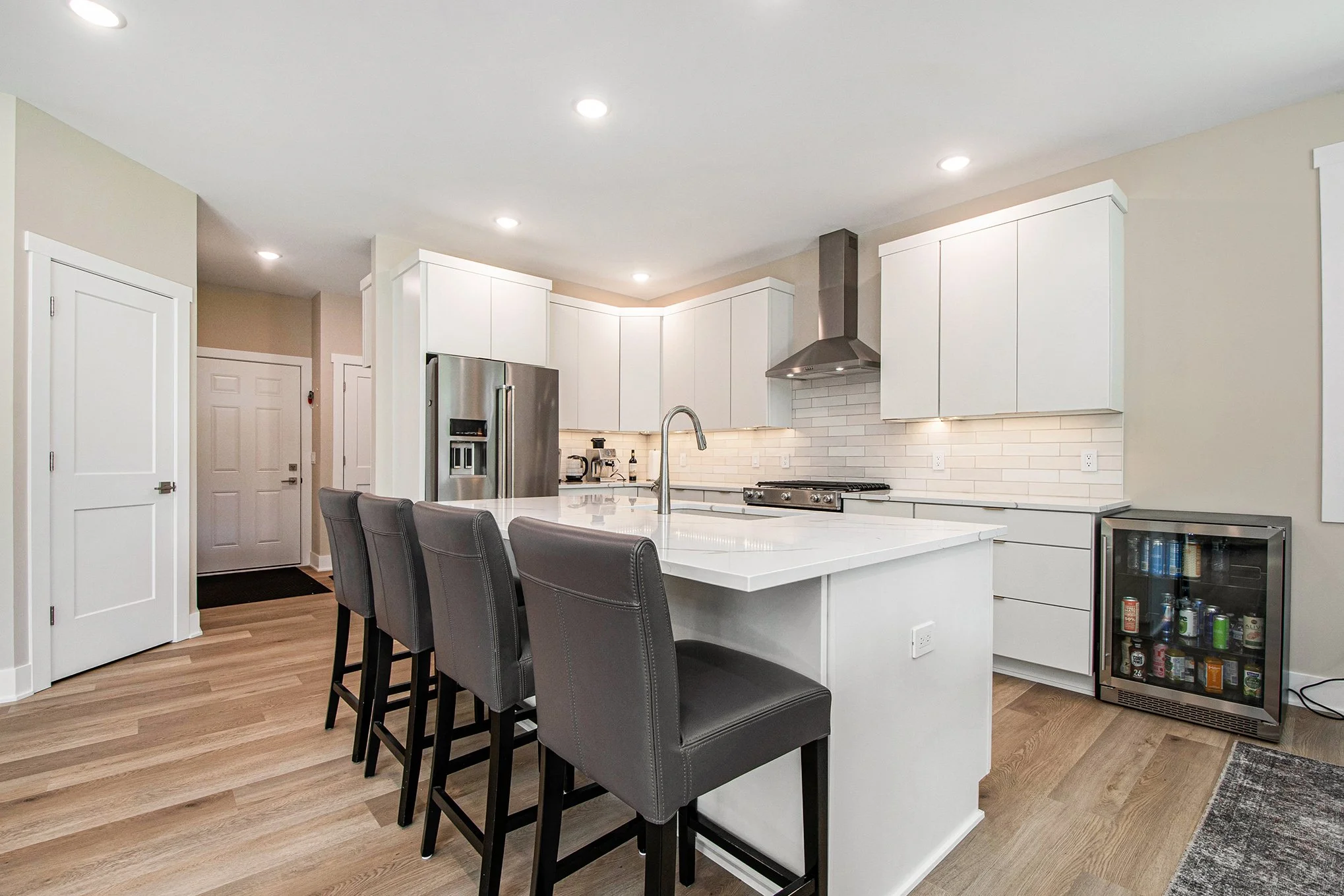 Modern kitchen with white cabinets, stainless steel refrigerator, island with sink and four gray barstools, white subway tile backsplash, and small beverage fridge.
