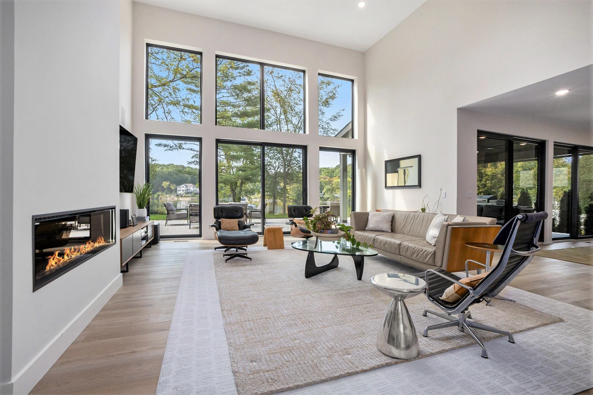 Living room with large windows showing trees and water outside, modern furniture including a beige sofa, black chairs, glass coffee table, fireplace, and a patio area with outdoor seating.