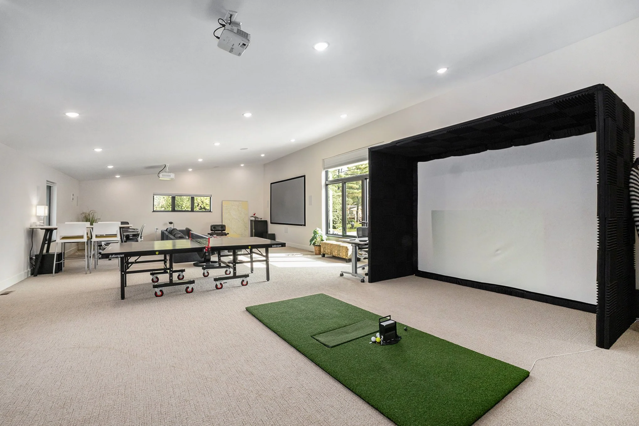 Spacious basement with a ping pong table, a golf simulator with a green mat, a black sofa, a wall-mounted TV, and a small table with chairs, illuminated by ceiling lights, with a window bringing in natural light.